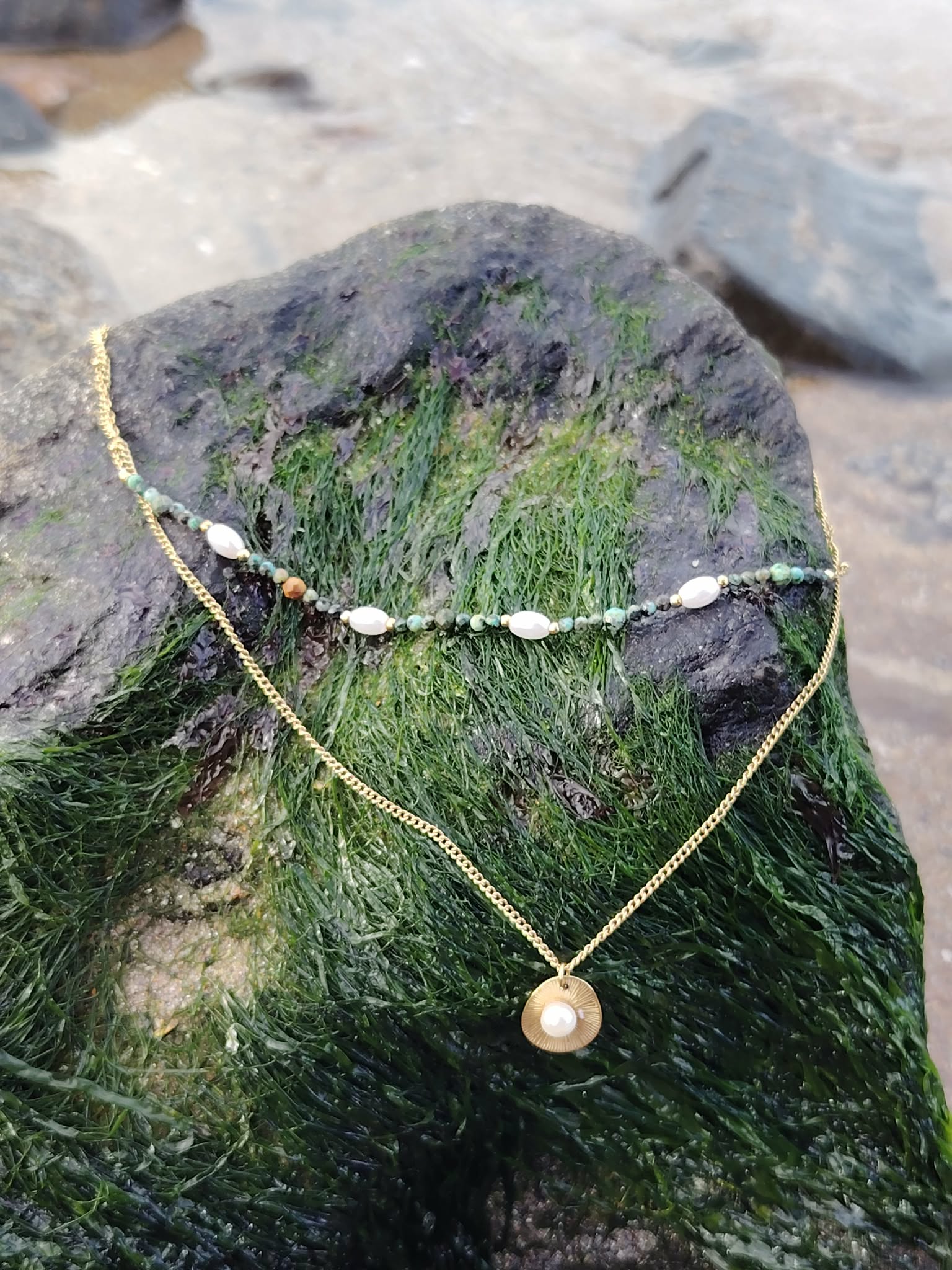 Collier double chaîne dorée en acier inoxydable avec perles blanches, pierres vertes et médaillon nacré, posé sur une souche de bois recouverte de mousse verte, ambiance nature, lumière douce en Bretagne