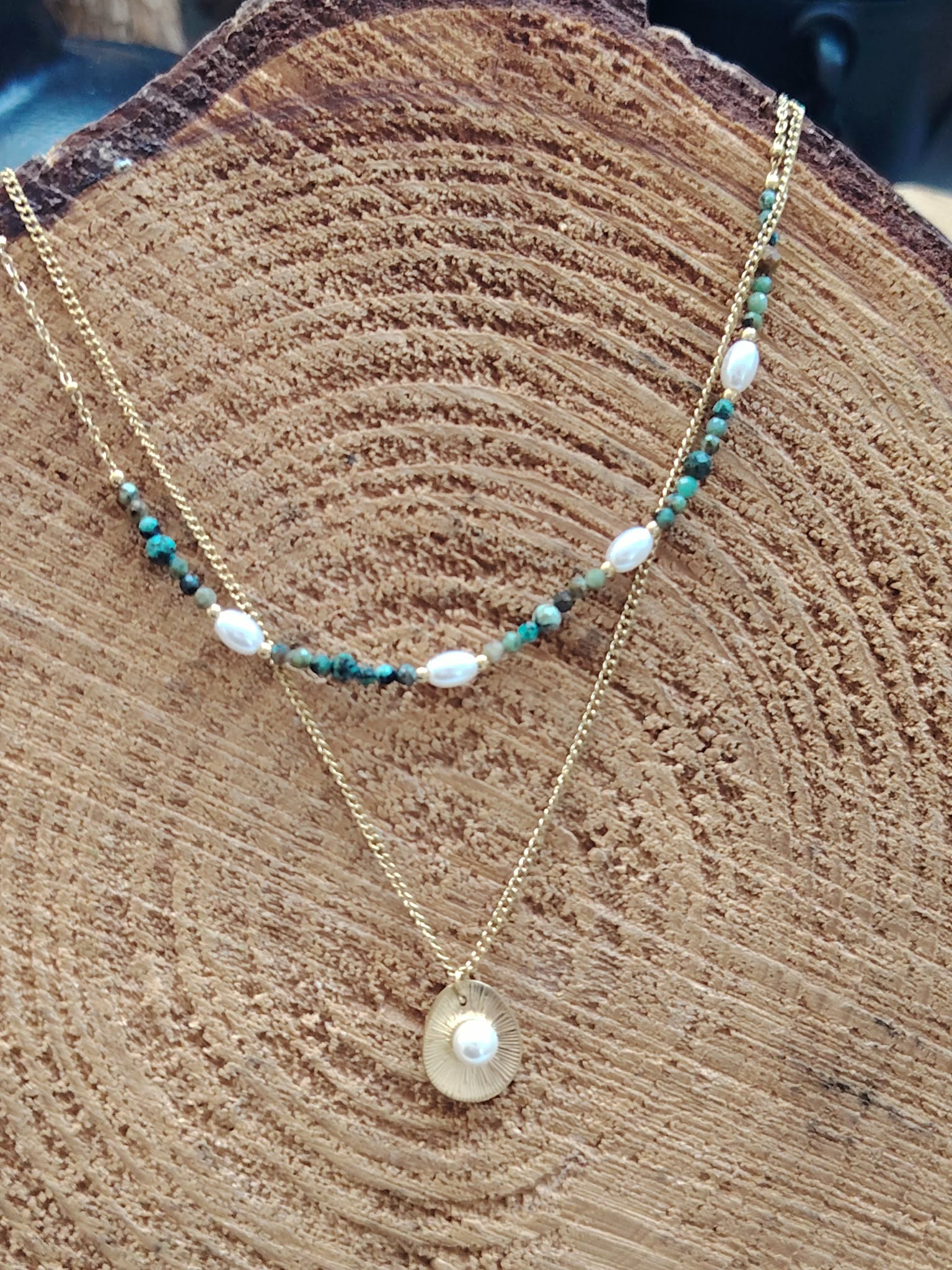 Collier double chaîne dorée acier inoxydable, perles blanches, pierres vertes, médaillon nacré, sur tranche de bois.