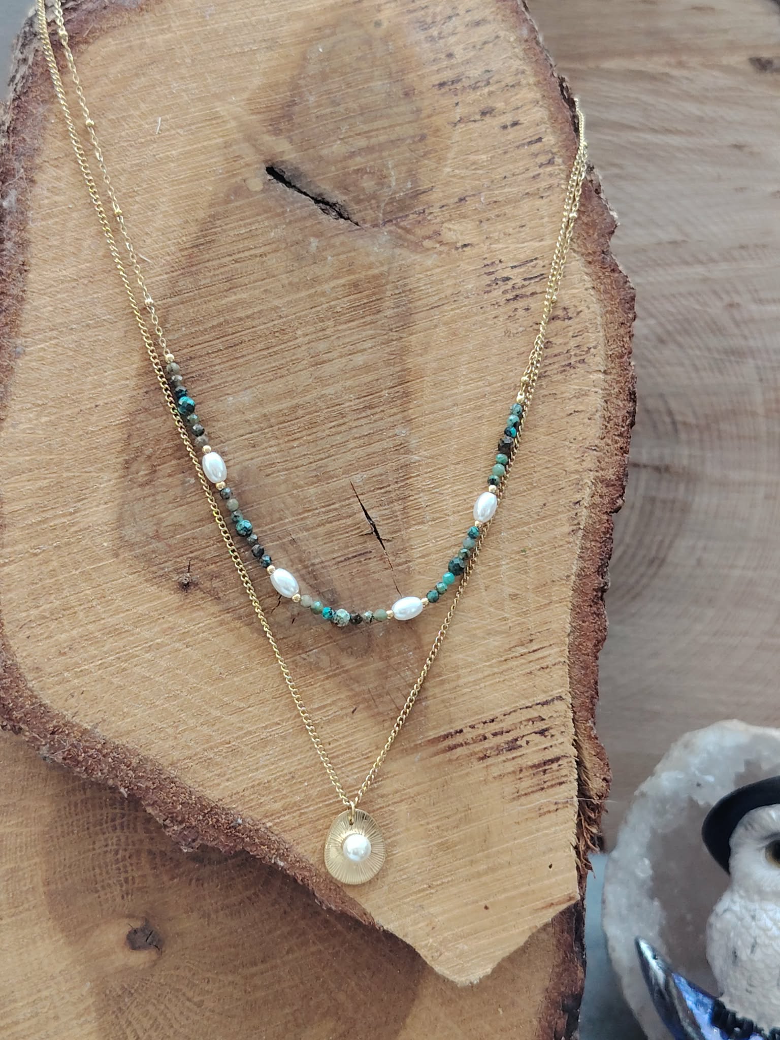 Collier double chaîne dorée acier inoxydable, perles blanches, pierres vertes, médaillon nacré, présenté sur buste en bois.