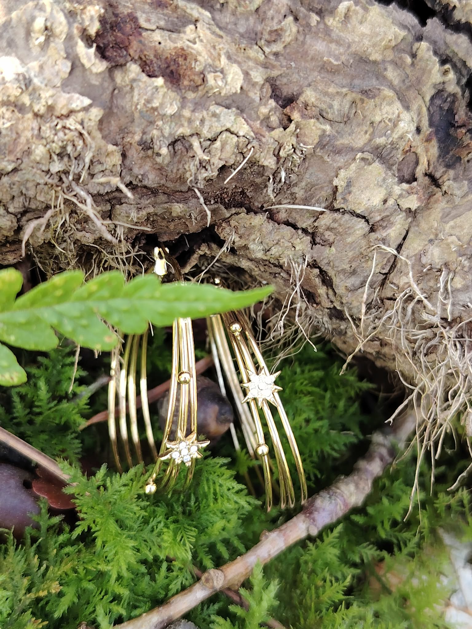Boucles d’oreilles dorées 4 anneaux ovales reliés avec étoiles strassées, posées en décor nature sur mousse verte au pied d’une écorce, avec feuille et petite branche au premier plan.
