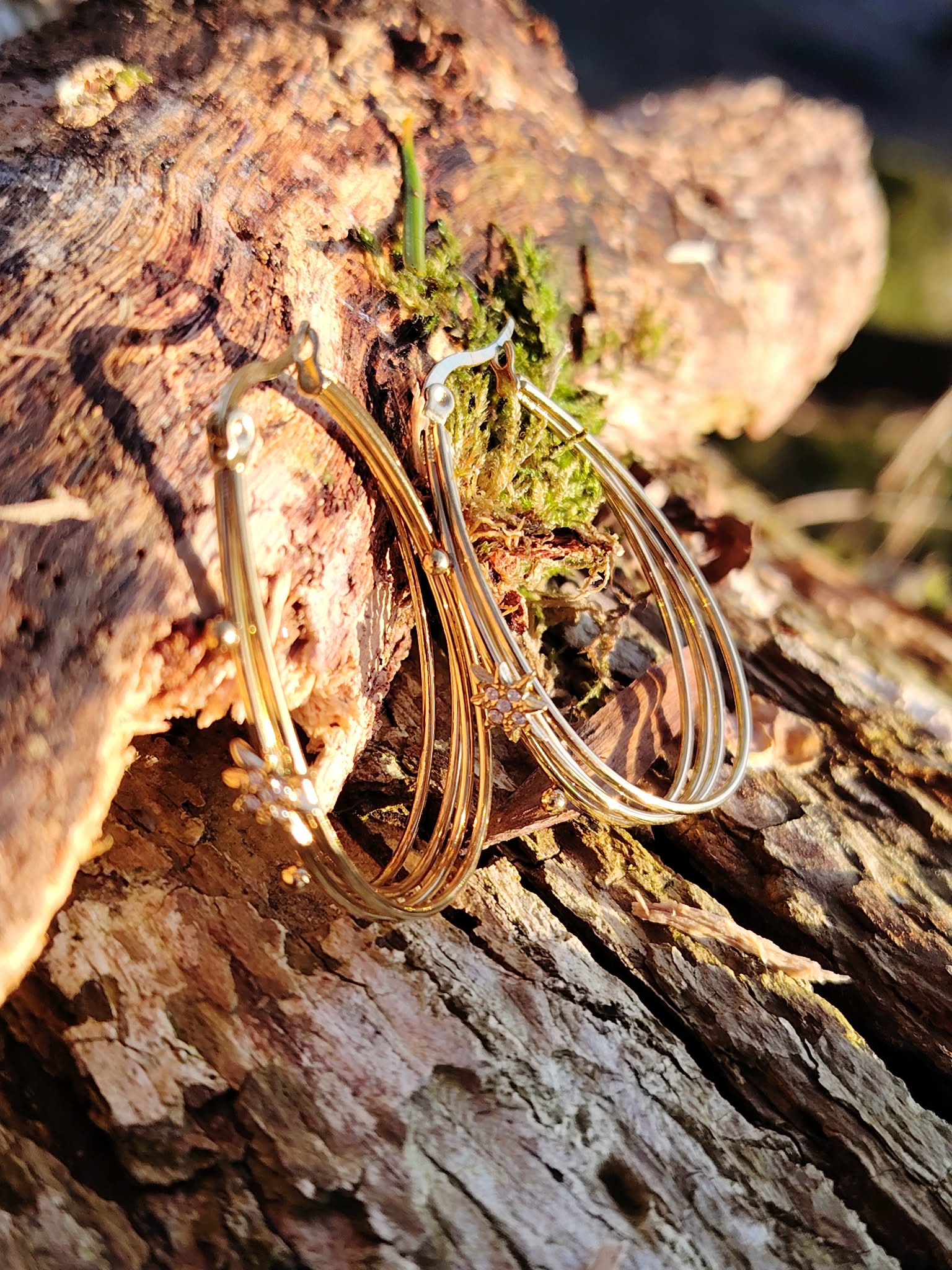 Boucles d’oreilles dorées 4 anneaux ovales reliés avec étoiles strassées, posées en décor nature dans un creux d’écorce/bois brut, avec mousse et petits brins végétaux, en lumière chaude.