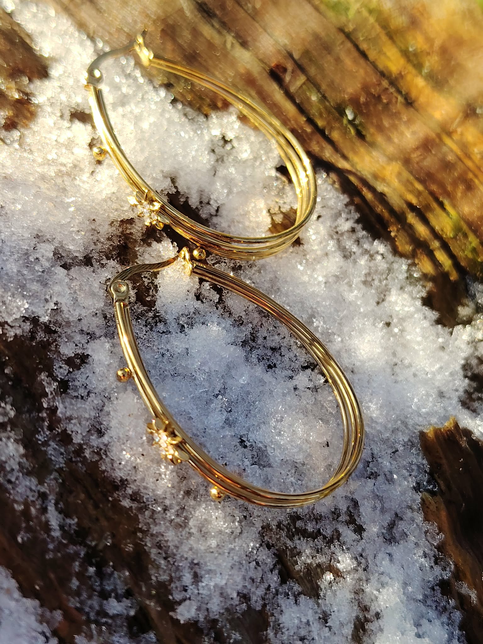 Boucles d’oreilles dorées à 4 anneaux ovales reliés, étoiles strassées, posées sur neige fondue avec décor bois en arrière-plan. en Bretagne