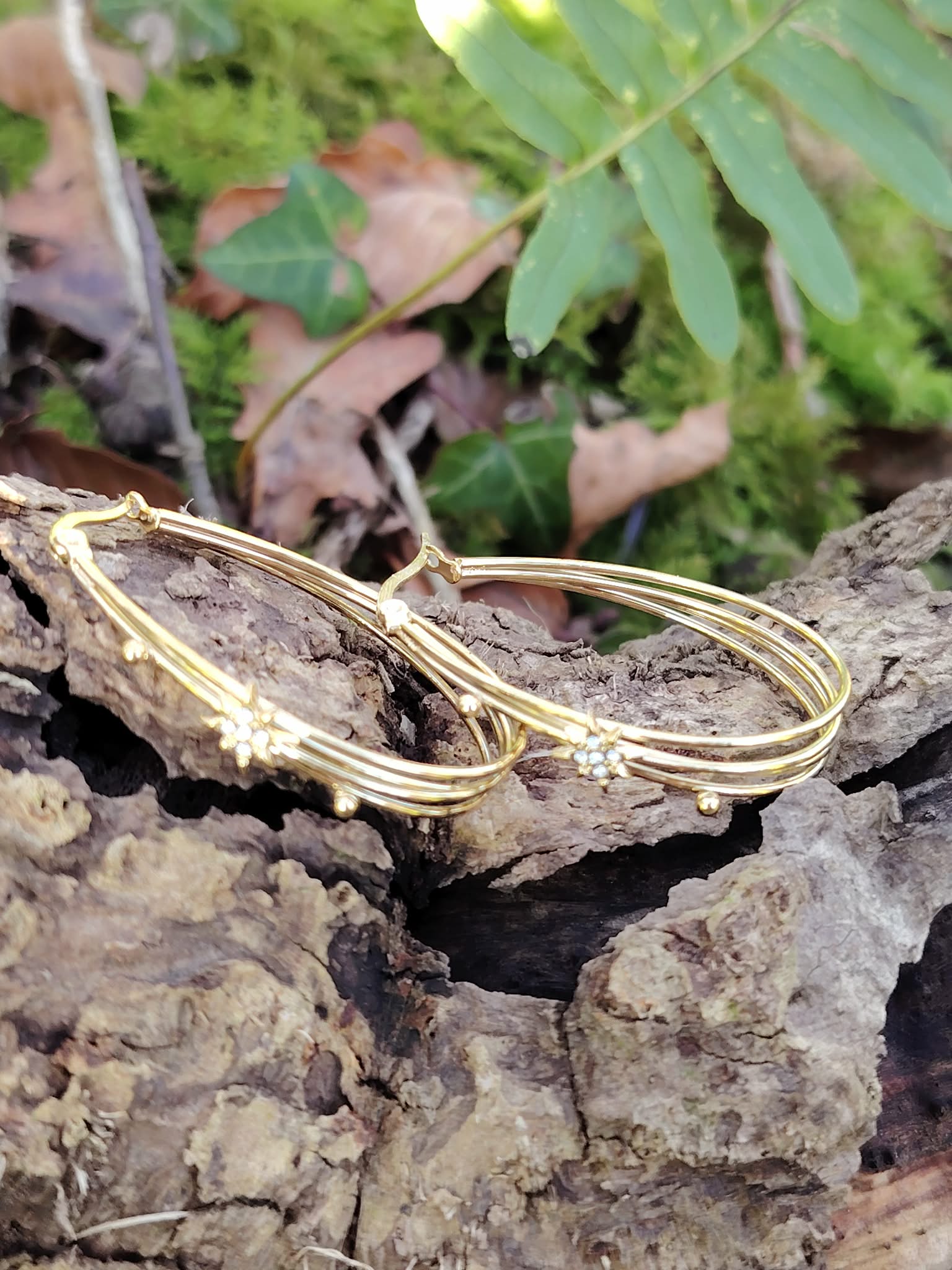 Boucles d’oreilles dorées 4 anneaux ovales reliés, détail étoile strassée, posées sur écorce avec mousse verte et feuilles en décor naturel en Bretagne