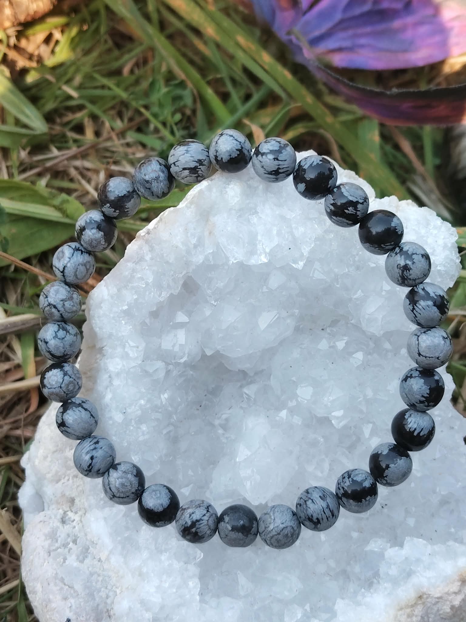 Bracelet obsidienne neige 6 mm posé sur galet clair, perles noir profond et gris nuage, feuillage en arrière-plan, protection douce, ancrage, calme intérieur, pensées plus nettes.