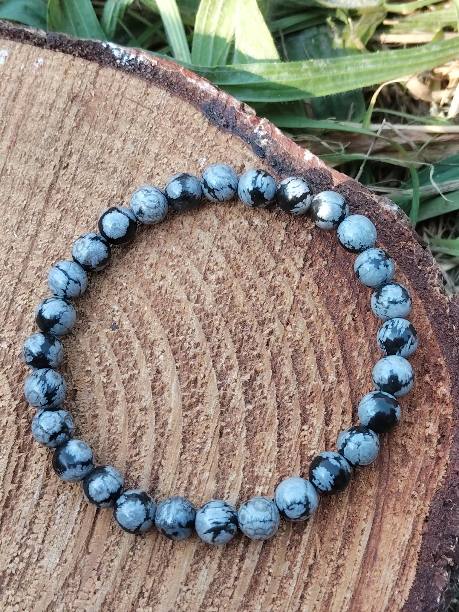 Bracelet obsidienne neige 6 mm porté au poignet, perles noir profond et gris moucheté, photo en extérieur avec feuillage, stabilité, protection, clarté mentale, recentrage.