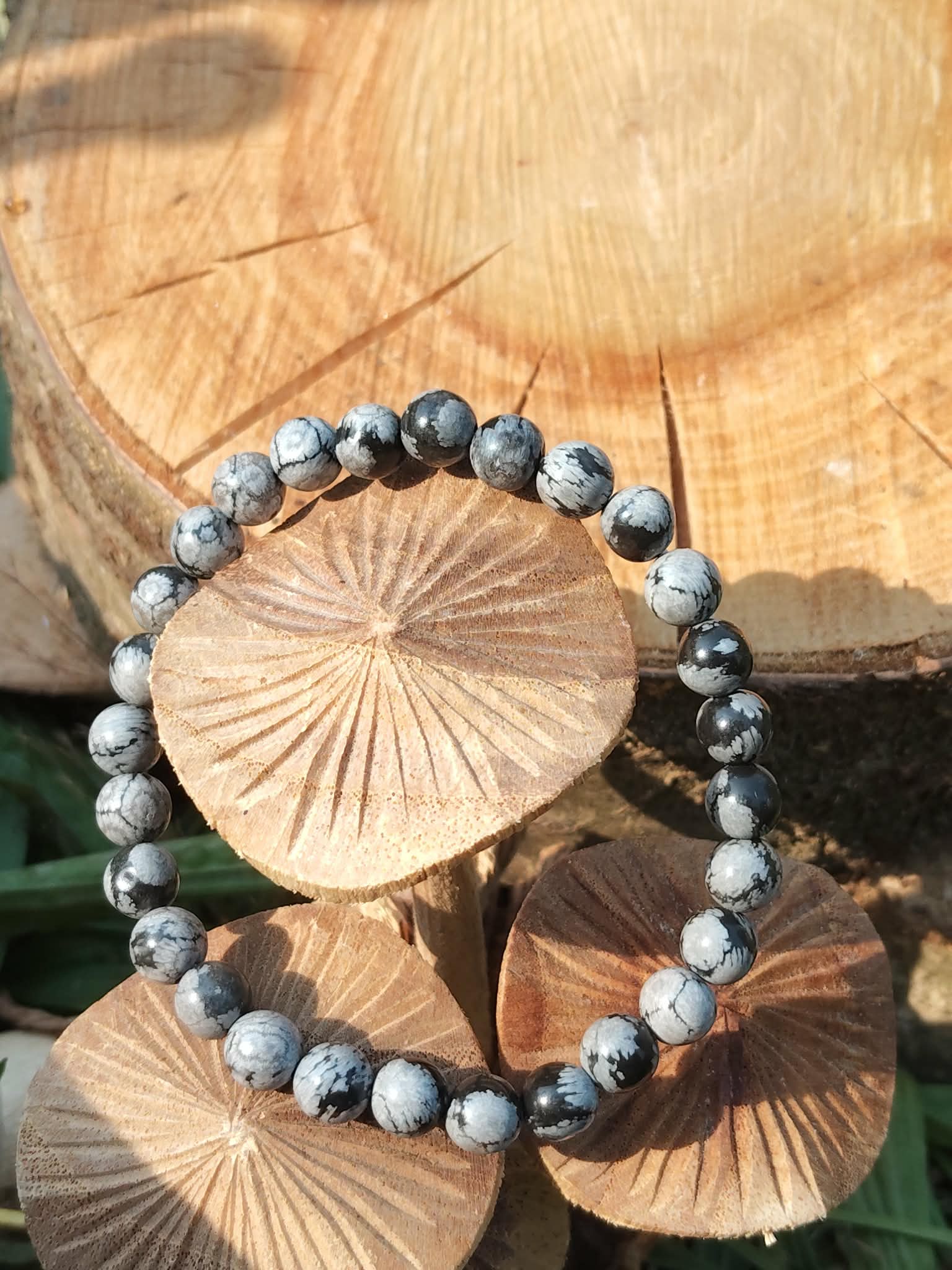 Bracelet obsidienne neige 6 mm posé sur galet clair avec feuillage en arrière-plan, perles noir profond et gris nuage pour clarté du mental et stabilité émotionnelle