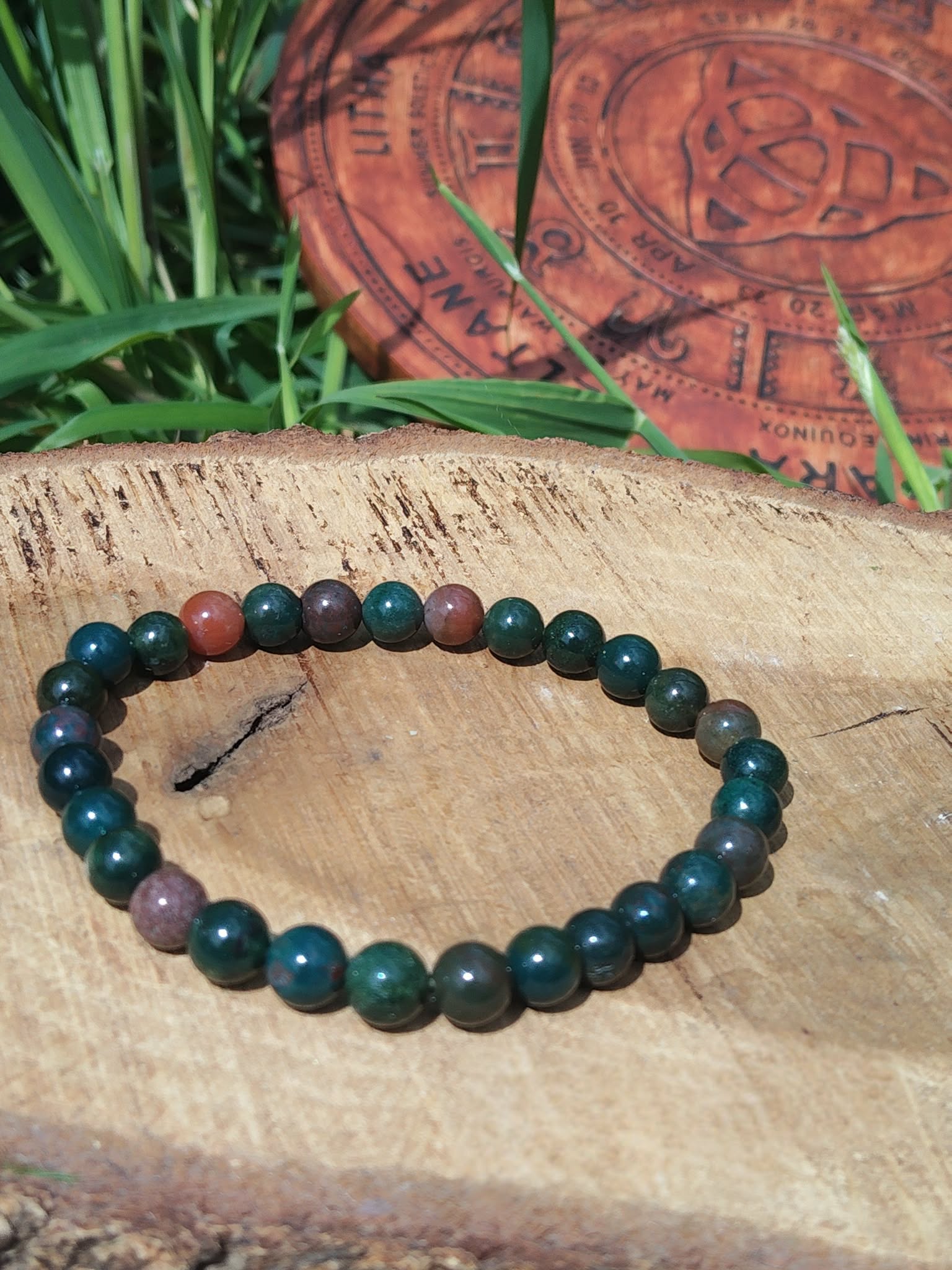 Bracelet jaspe héliotrope 6 mm sur rondin de bois, verdure en fond ; stabilité, apaisement, confiance.