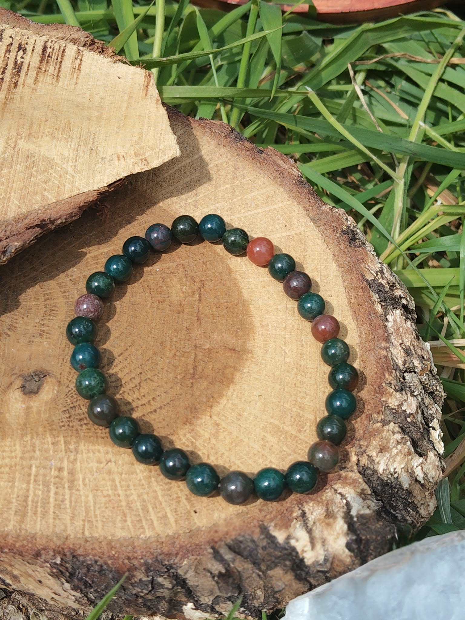 Bracelet jaspe héliotrope 6 mm sur rondin de bois, herbes en fond, lumière naturelle ; ancrage, calme, courage.