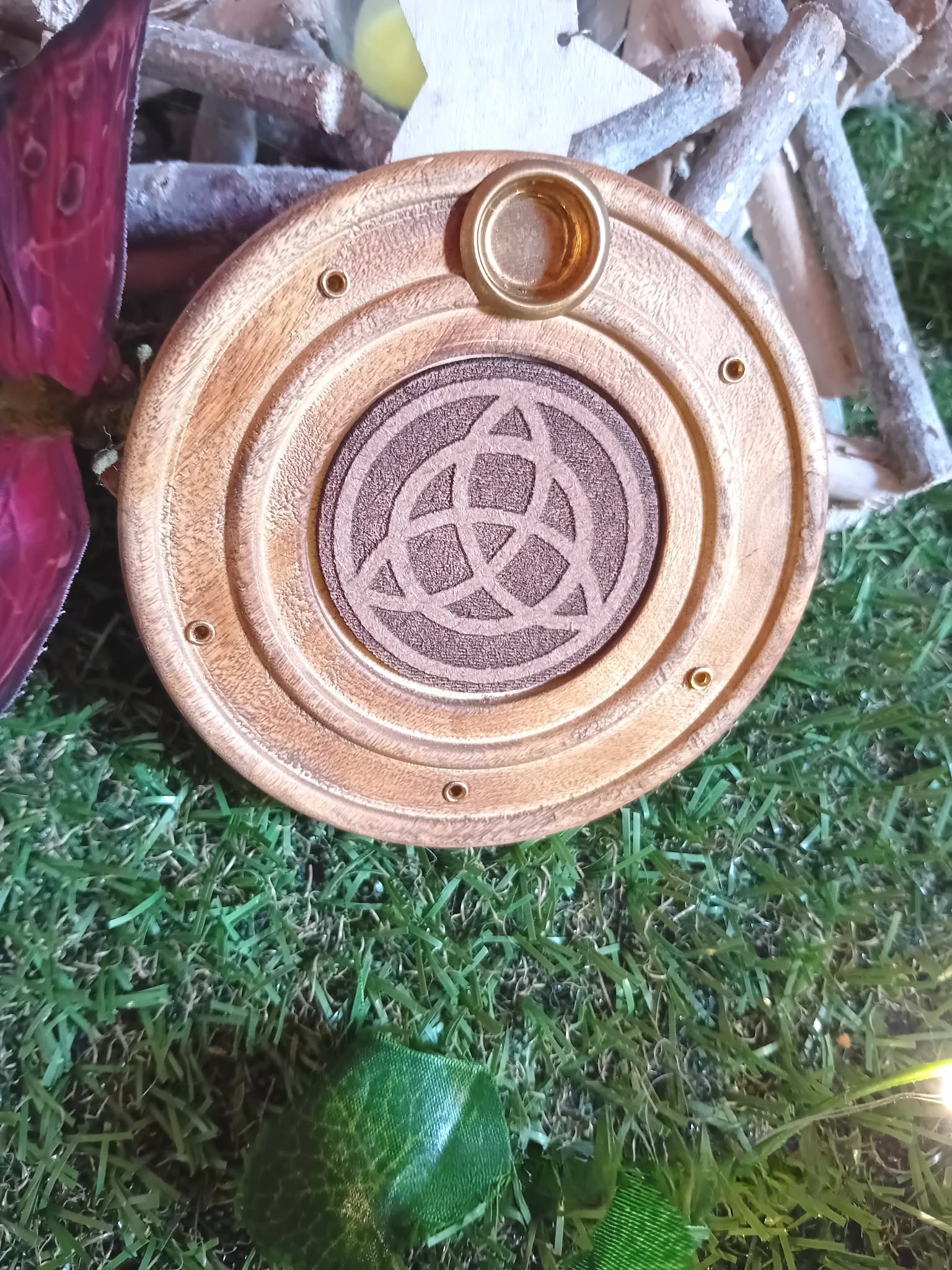 Porte-encens bois rond triquetra, posé sur mousse verte avec décor branches en bois et touche bordeaux en arrière-plan, ambiance nature.