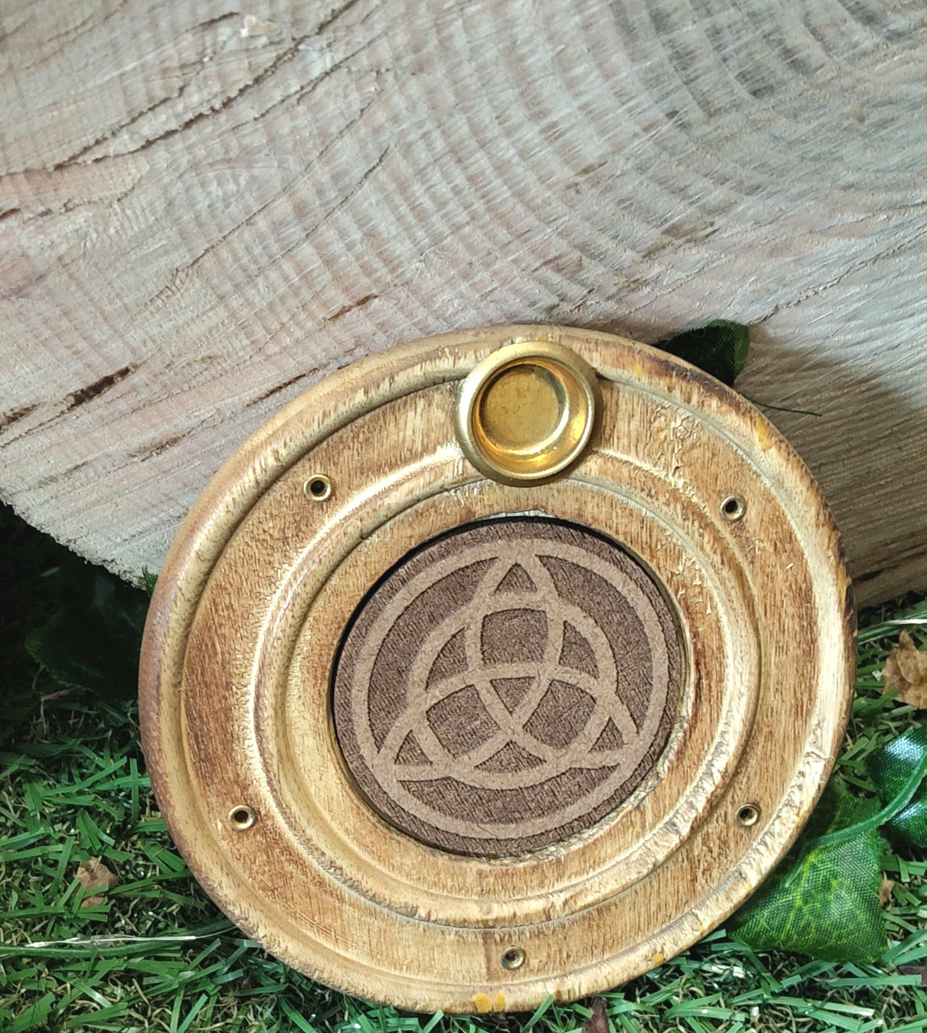 Porte-encens rond en bois motif triquetra, posé sur planche claire avec verdure en fond, ambiance protection, harmonie, rituel.