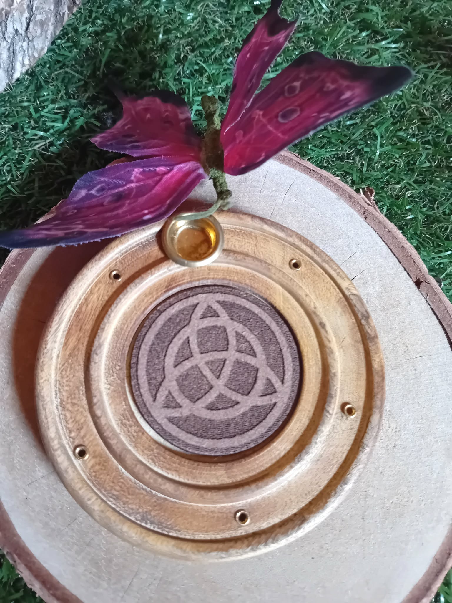 Porte-encens bois rond triquetra, posé sur mousse verte avec papillon rose en premier plan, fond clair, ambiance douce.