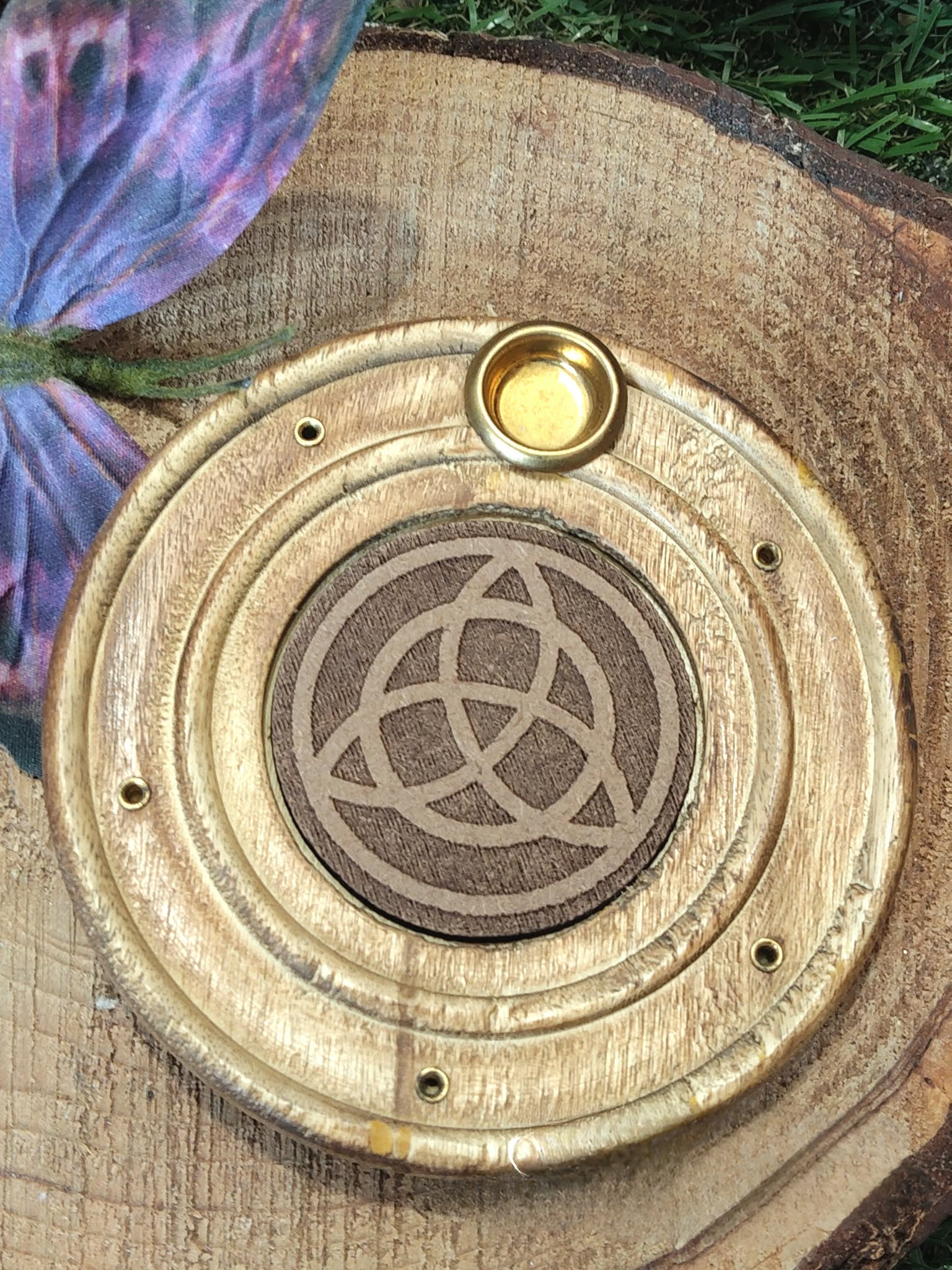 Porte-encens bois rond triquetra, posé sur tranche de bois brut avec papillon bleu-violet en décor, fond végétal sombre, ambiance calme.