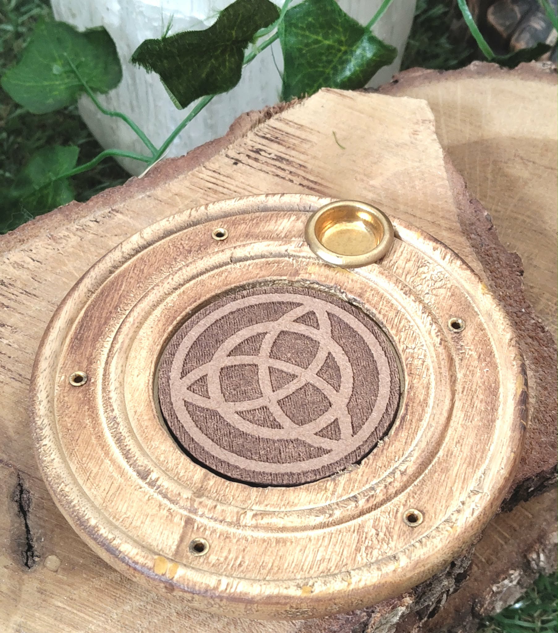 Porte-encens bois rond triquetra, posé sur tranche de bois brut avec feuillage vert en fond, ambiance calme et protection symbolique.