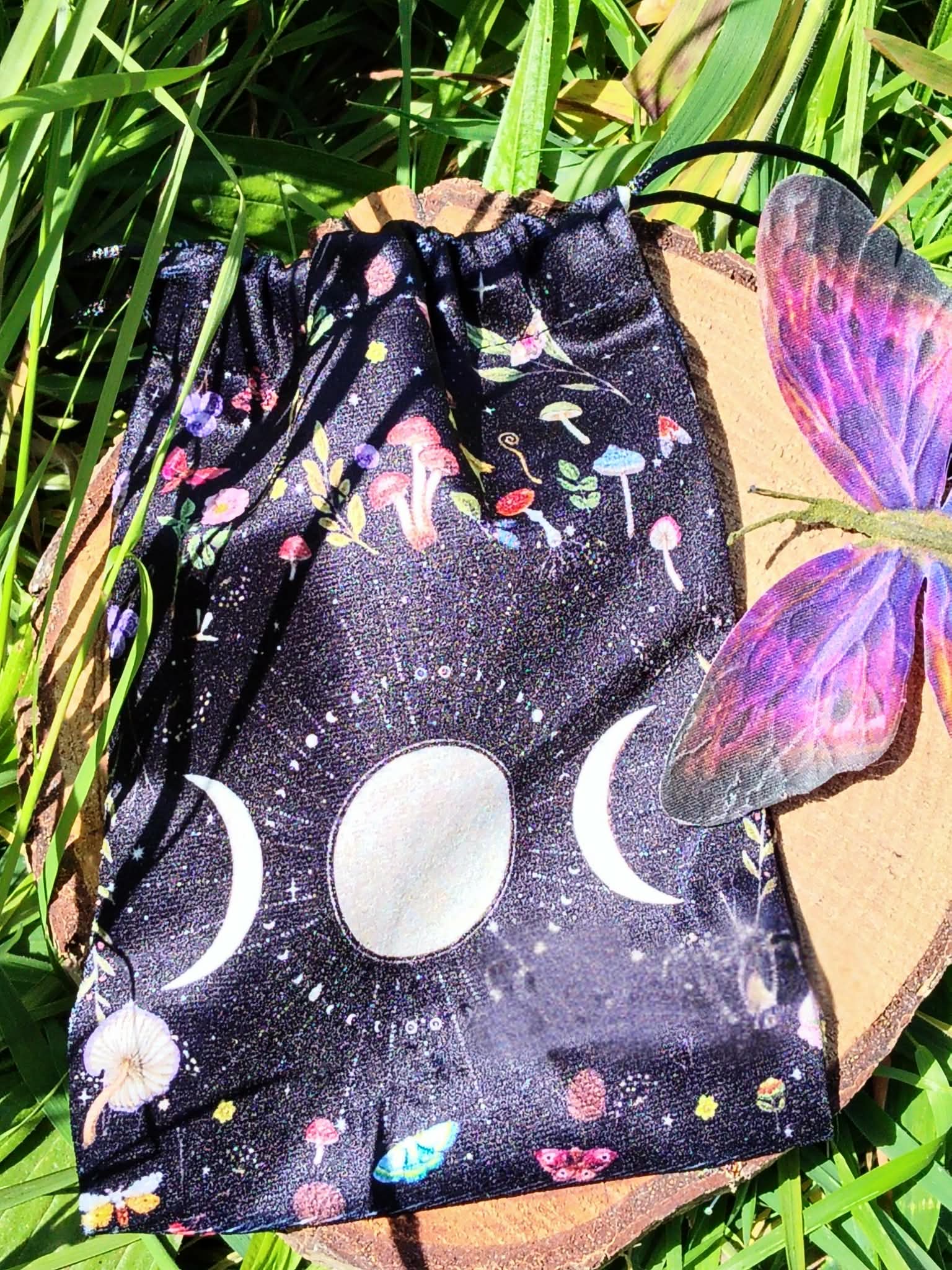 Pochon de rangement en velours noir, symbole triple lune blanc (croissant, pleine lune, croissant) sur l’avant, cordons de serrage en haut, posé sur planche de bois, entouré de feuillage vert, lumière naturelle, ambiance mystique et naturelle.