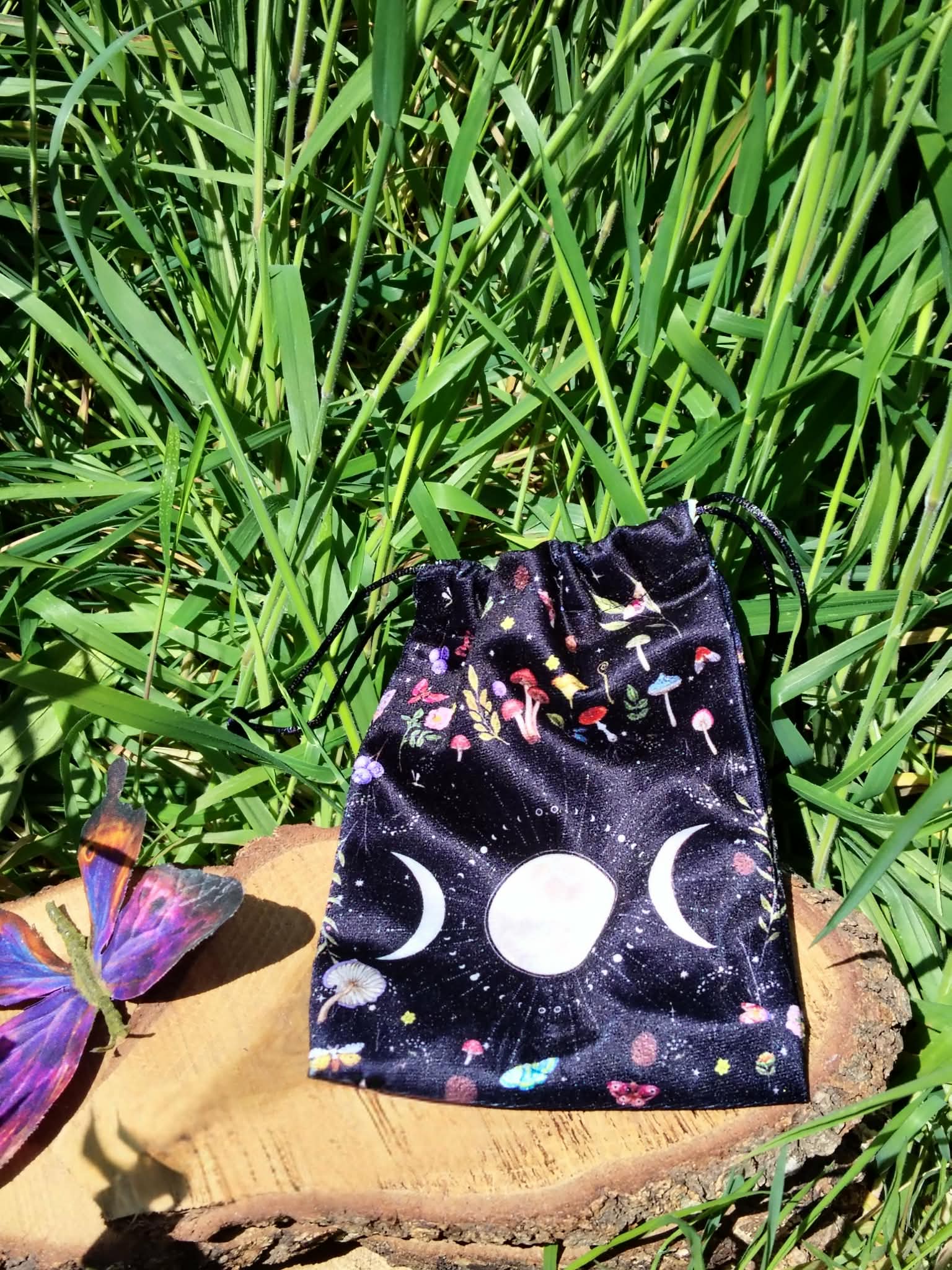 Pochon de rangement en velours noir motif galaxie, symbole triple lune blanc sur l’avant, présenté sur rondin de bois avec feuillage vert en arrière-plan, papillon décoratif violet sur le côté, lumière naturelle, ambiance nature et mystique.