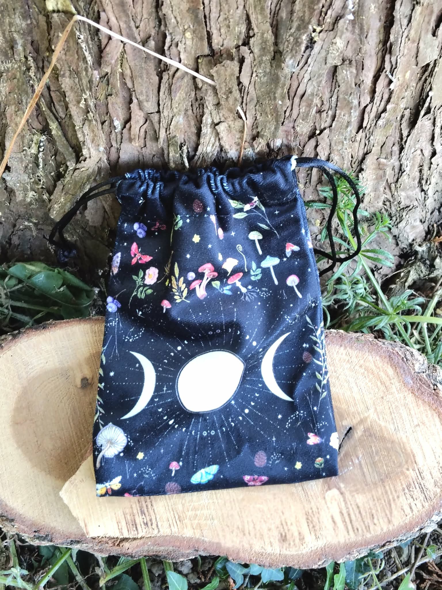 Pochon de rangement en velours noir motif ciel étoilé, symbole triple lune blanc sur l’avant, présenté sur rondin de bois devant une écorce d’arbre, avec feuillage vert en décor, lumière naturelle, ambiance nature et ésotérique.