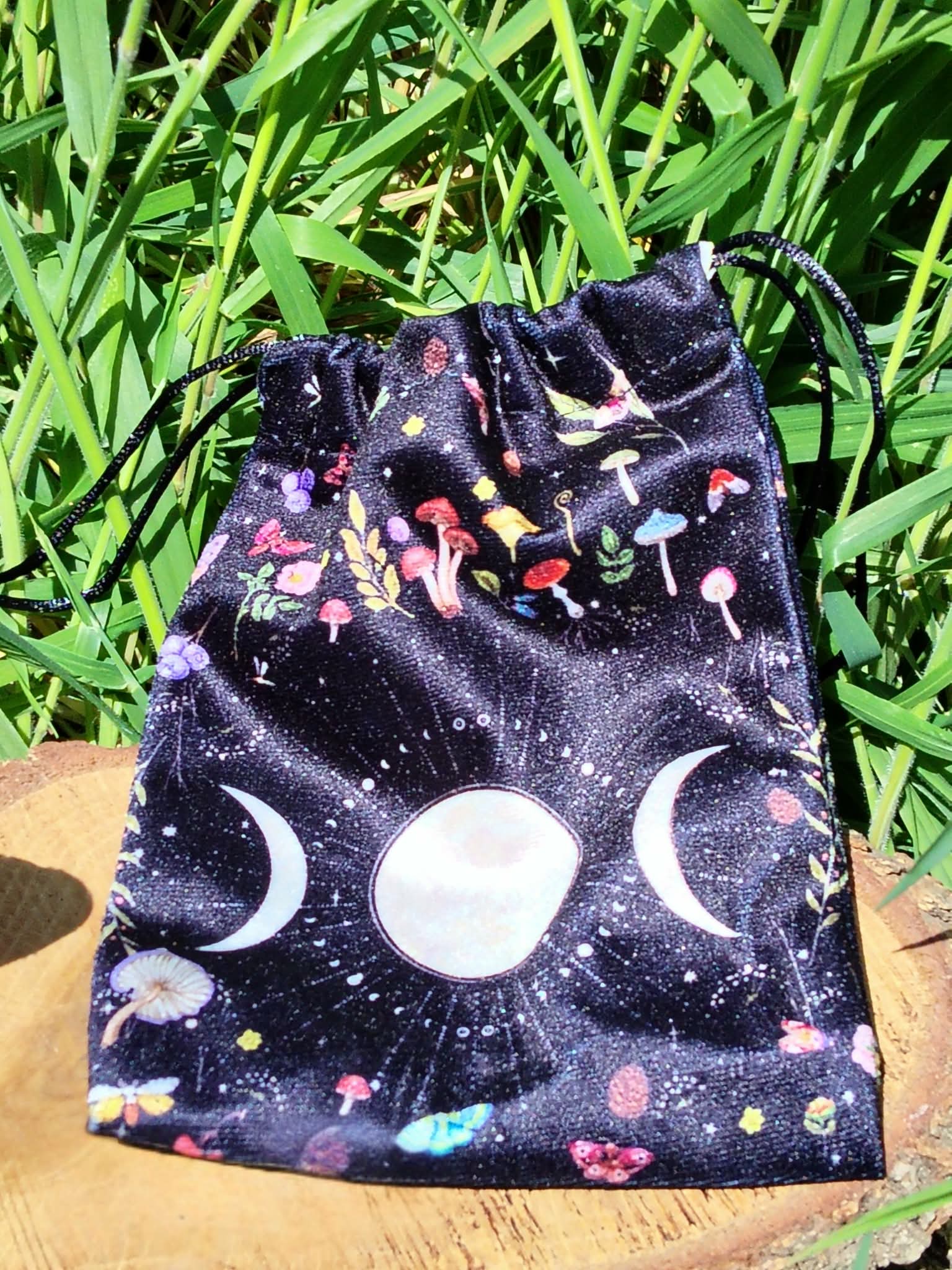 Pochon de rangement en velours noir motif galaxie, symbole triple lune blanc sur l’avant, cordon de serrage visible, posé sur planche de bois avec grandes feuilles vertes en arrière-plan, lumière naturelle, ambiance ésotérique et nature