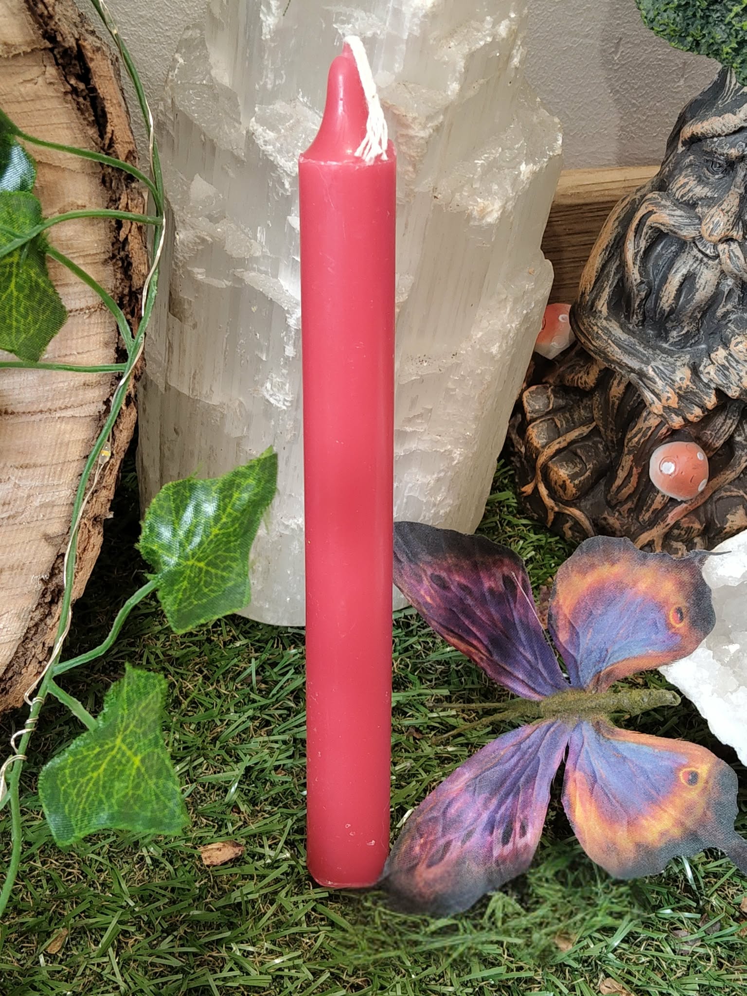 Bougie rouge fine plantée dans la mousse verte, papillon décoratif violet et orange au pied, lierre sur le côté, décor naturel avec planche de bois et éléments forestiers, énergie, vitalité, courage, motivation, transformation.