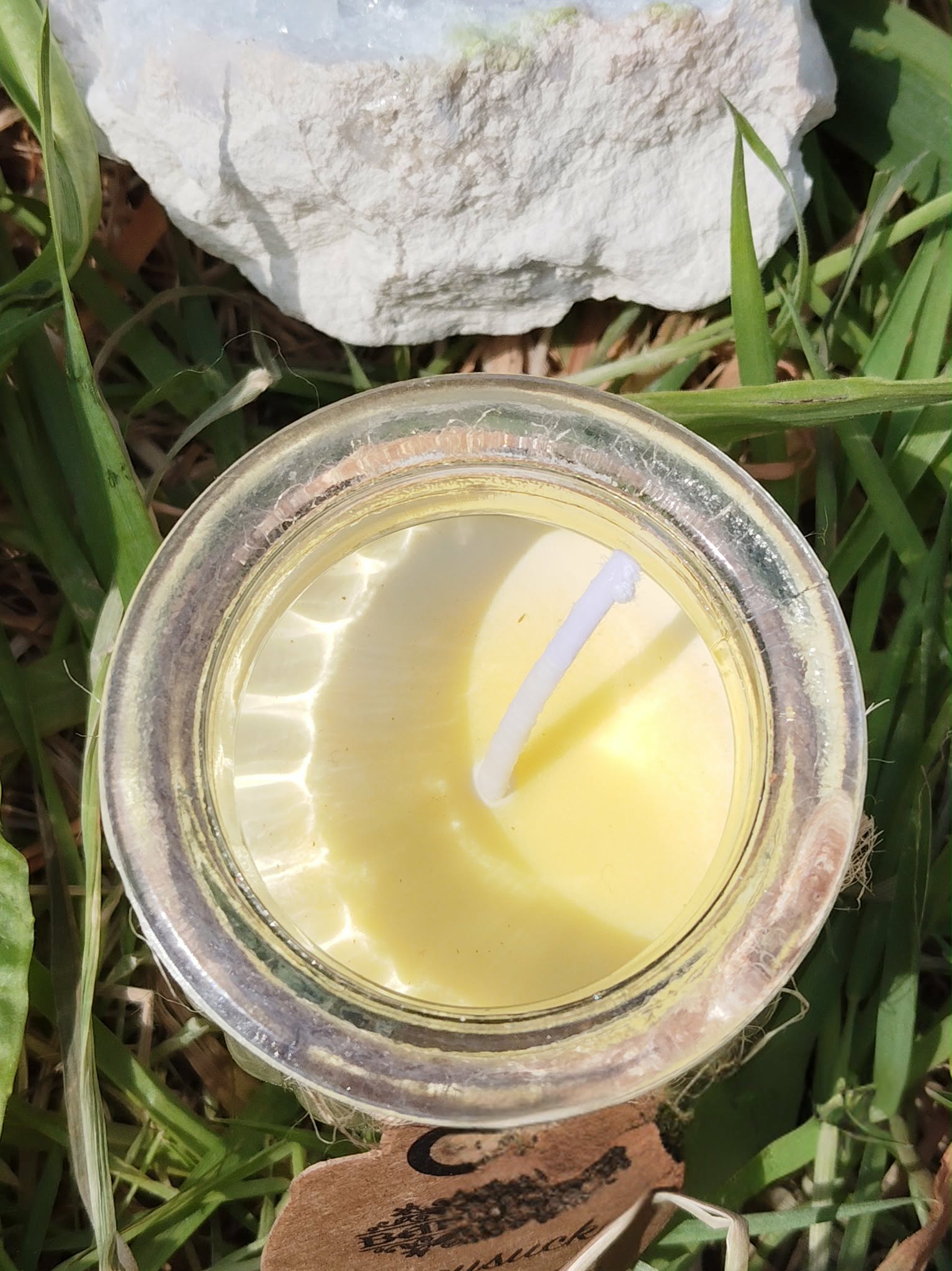 Vue du dessus d’une bougie en cire de soja couleur crème, mèche en coton au centre, dans un pot en verre, posée dans l’herbe avec amas de quartz en arrière-plan, ambiance nature, clarté, purification, apaisement, recentrage, douceur.