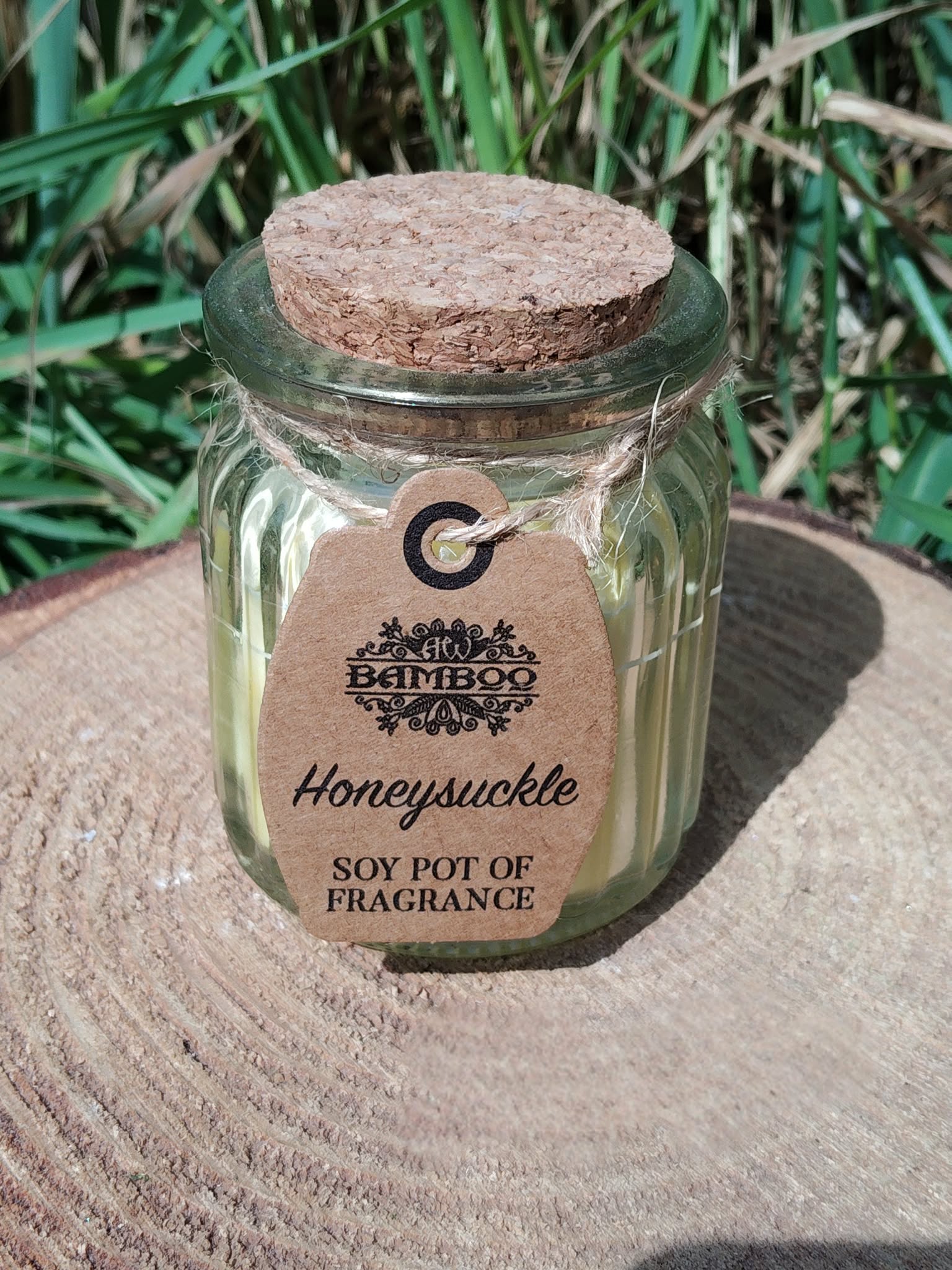 Bougie parfumée au chèvrefeuille en cire de soja, en pot en verre avec couvercle en liège, posée sur tissu beige avec feuillage vert en arrière-plan, ambiance naturelle, douceur, réconfort, apaisement, harmonie, cocooning.