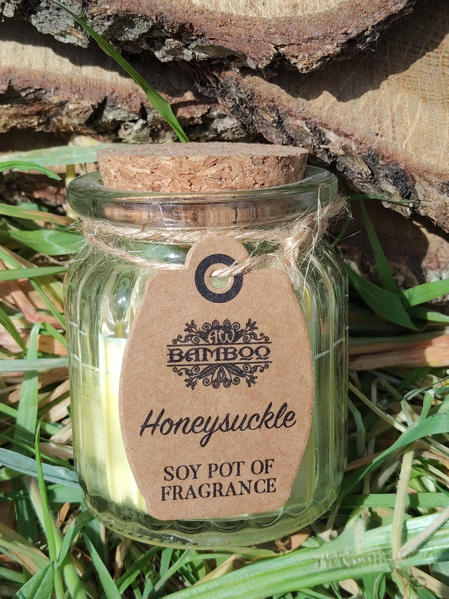 Bougie parfumée chèvrefeuille (Honeysuckle) en cire de soja, pot en verre à couvercle liège, posée dans l’herbe avec écorce en fond, ambiance nature, douceur, détente, réconfort, sérénité, chaleur du foyer.