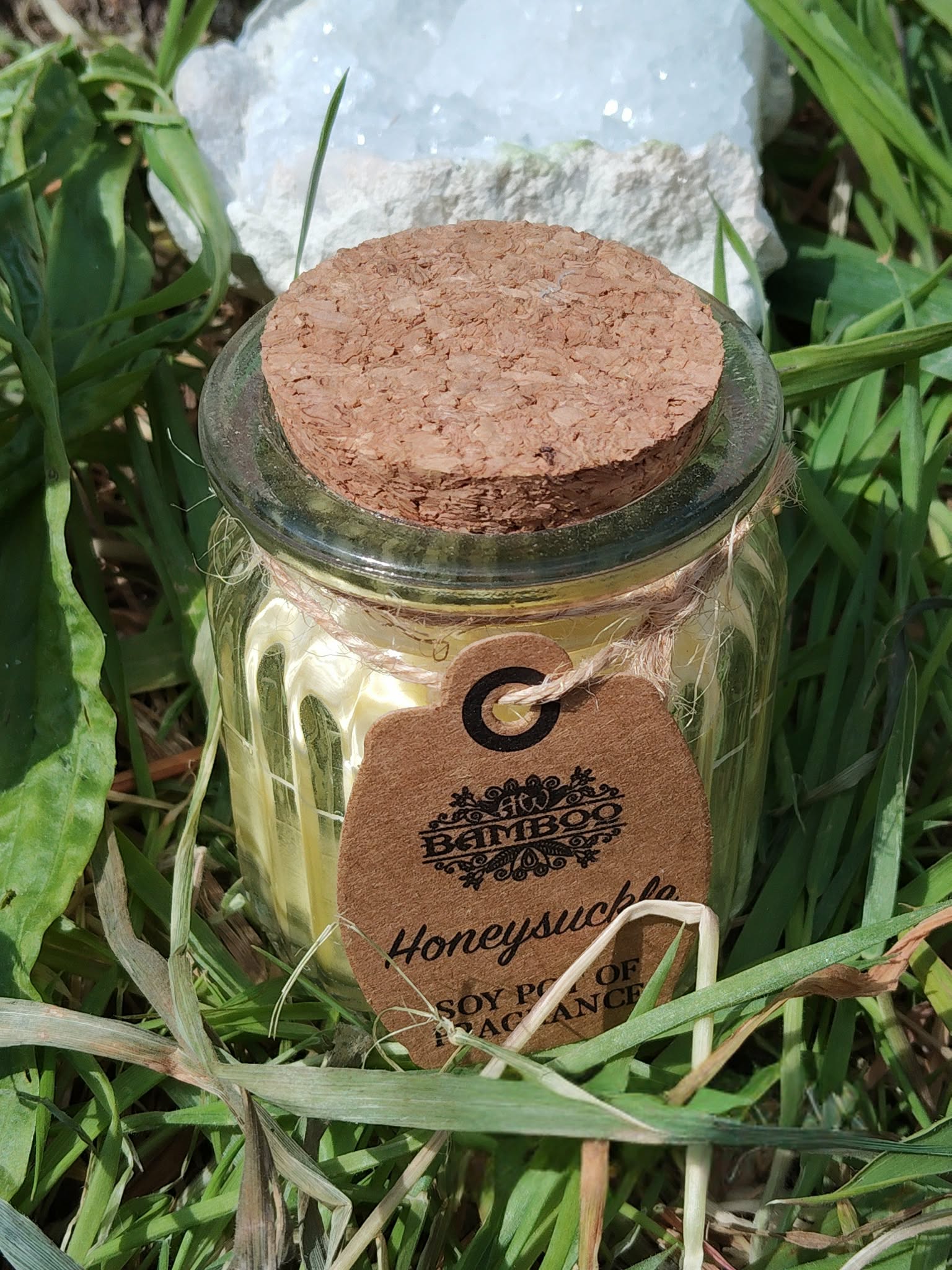 Bougie parfumée chèvrefeuille (Honeysuckle) en cire de soja, pot en verre avec couvercle en liège, posée dans l’herbe, amas de quartz en arrière-plan, décor nature, purification, clarté, apaisement, réconfort, harmonie, ambiance cocooning.
