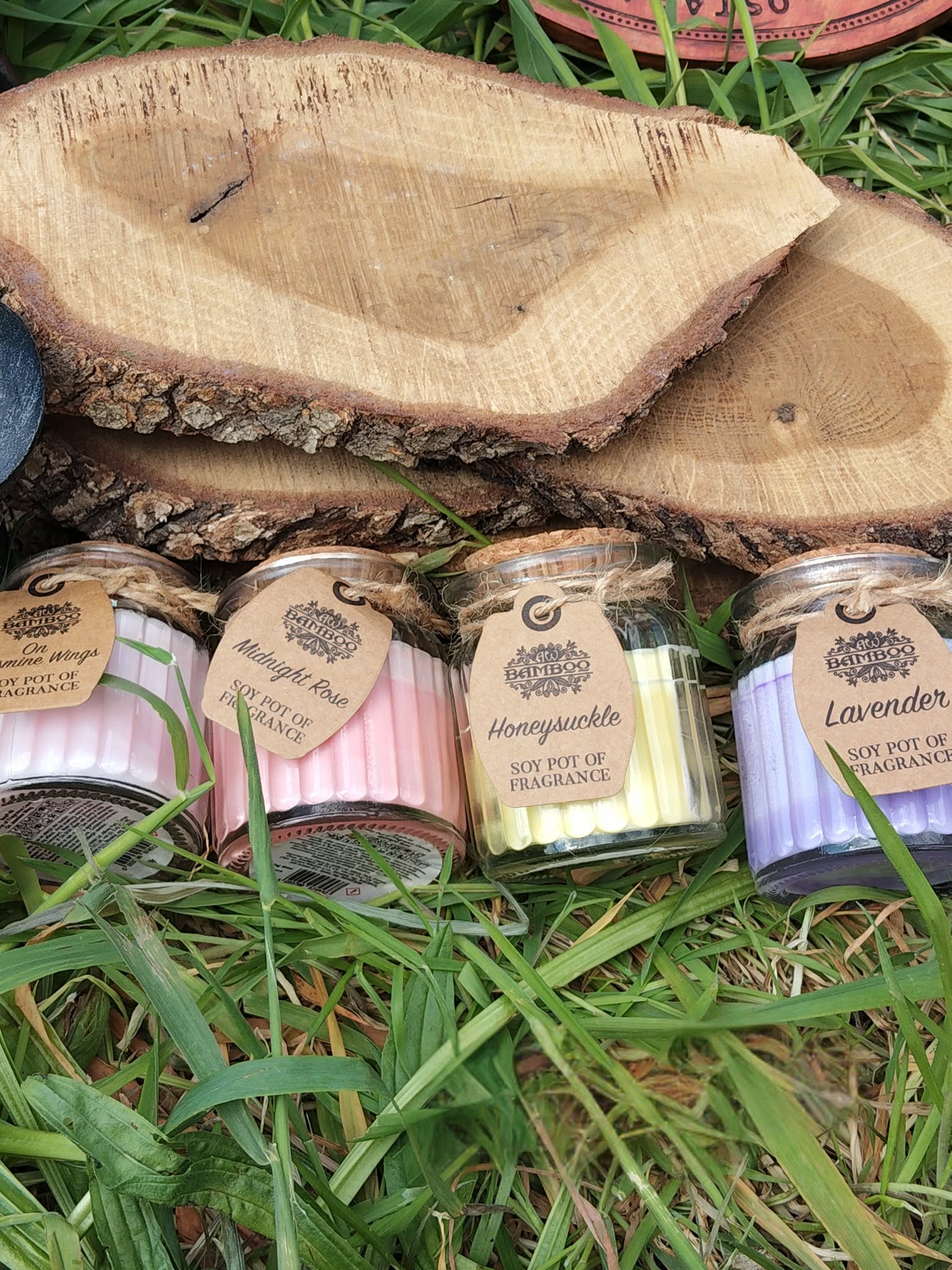 Bougies cire de soja en petits pots verre, couvercles liège, posées dans l’herbe au pied d’une tranche de bois, harmonie, détente, réconfort.