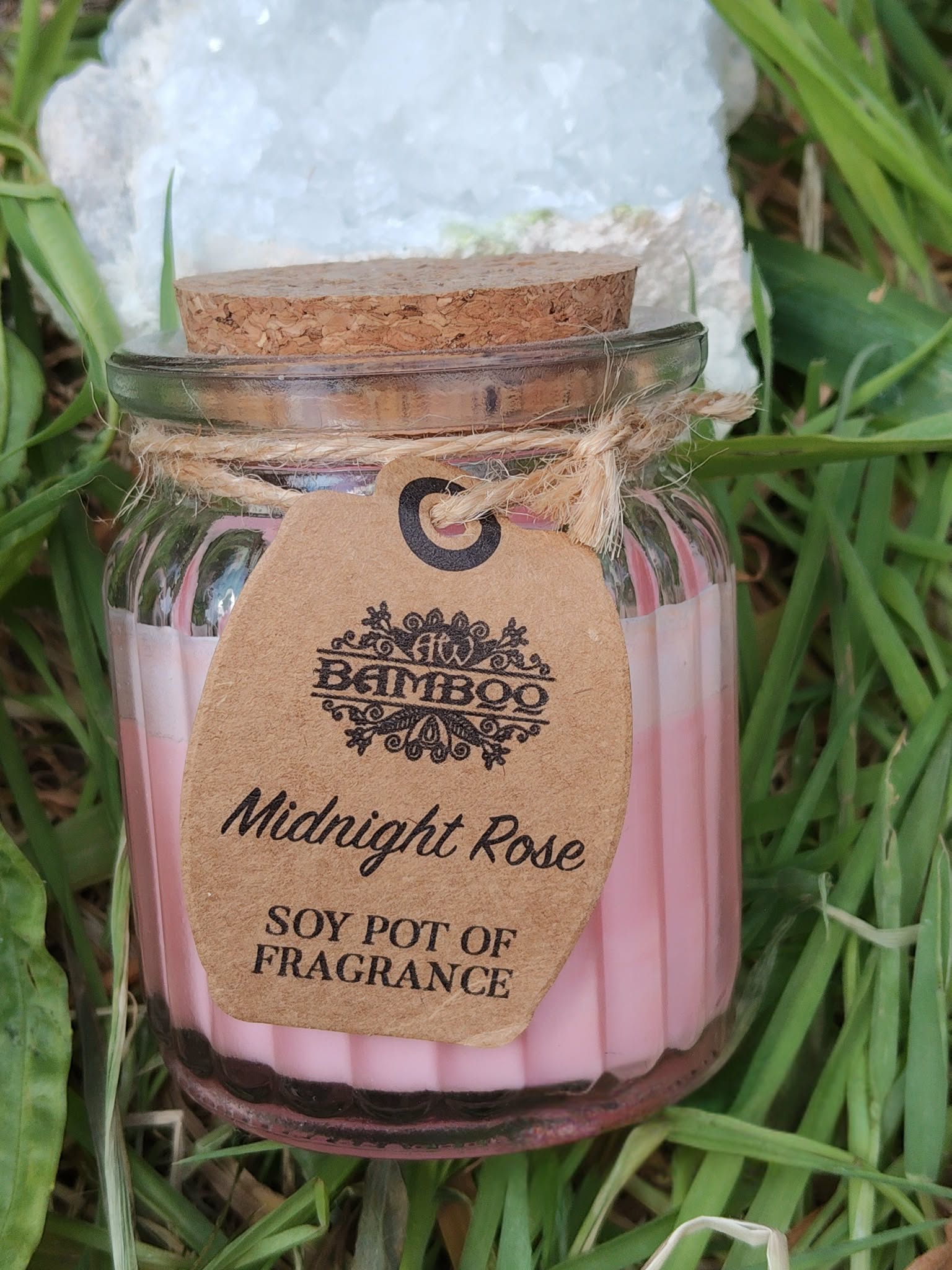 Bougie parfumée Rose de Minuit en cire de soja, pot en verre avec couvercle en liège et étiquette kraft, posée dans l’herbe en décor naturel, ambiance douce, détente, apaisement, cocooning.