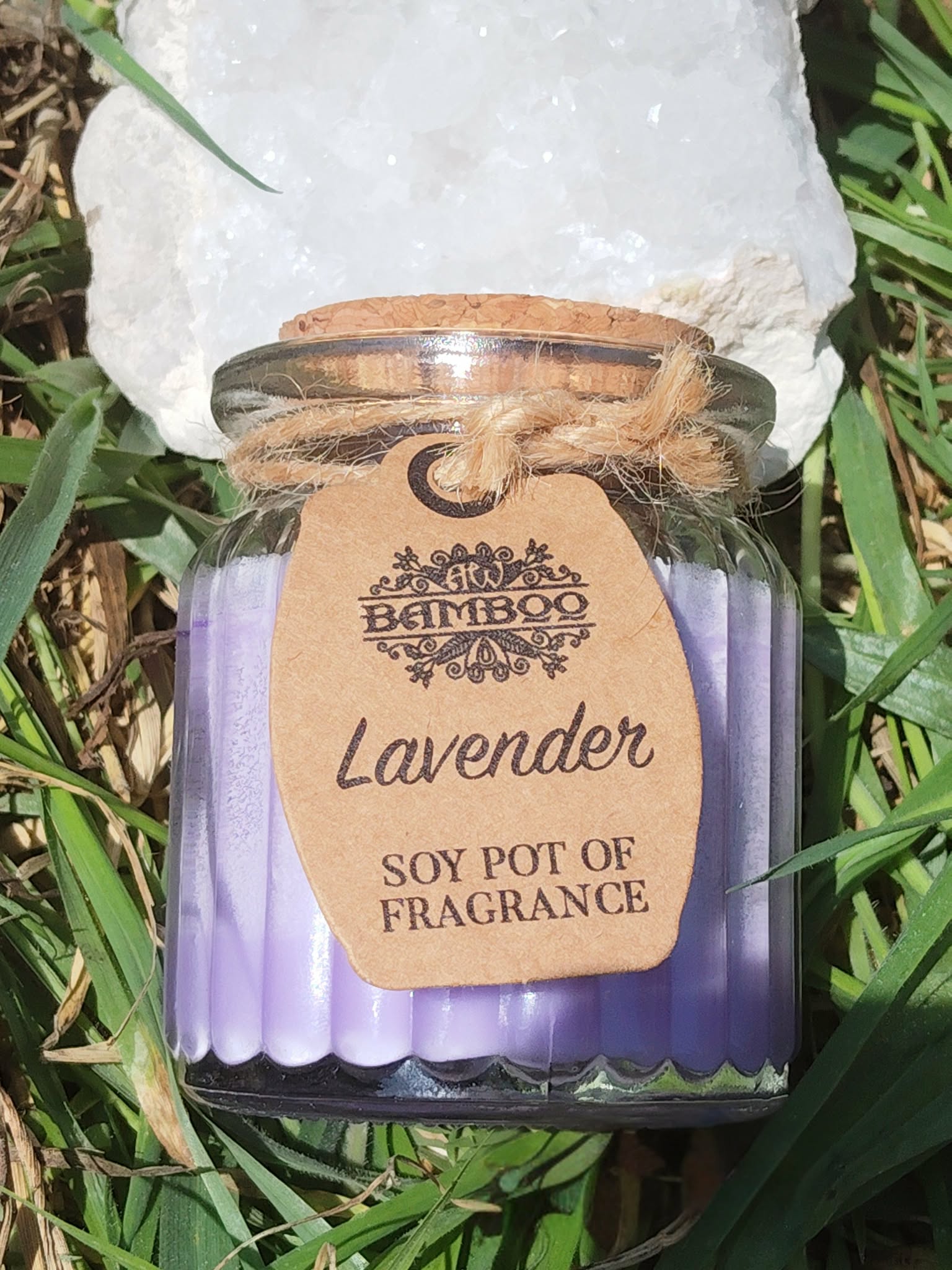 Bougie parfumée lavande en pot de verre strié violet avec couvercle en liège, ficelle et étiquette kraft, posée dans l’herbe en extérieur, lumière naturelle, ambiance douce, apaisante, détente, sérénité.