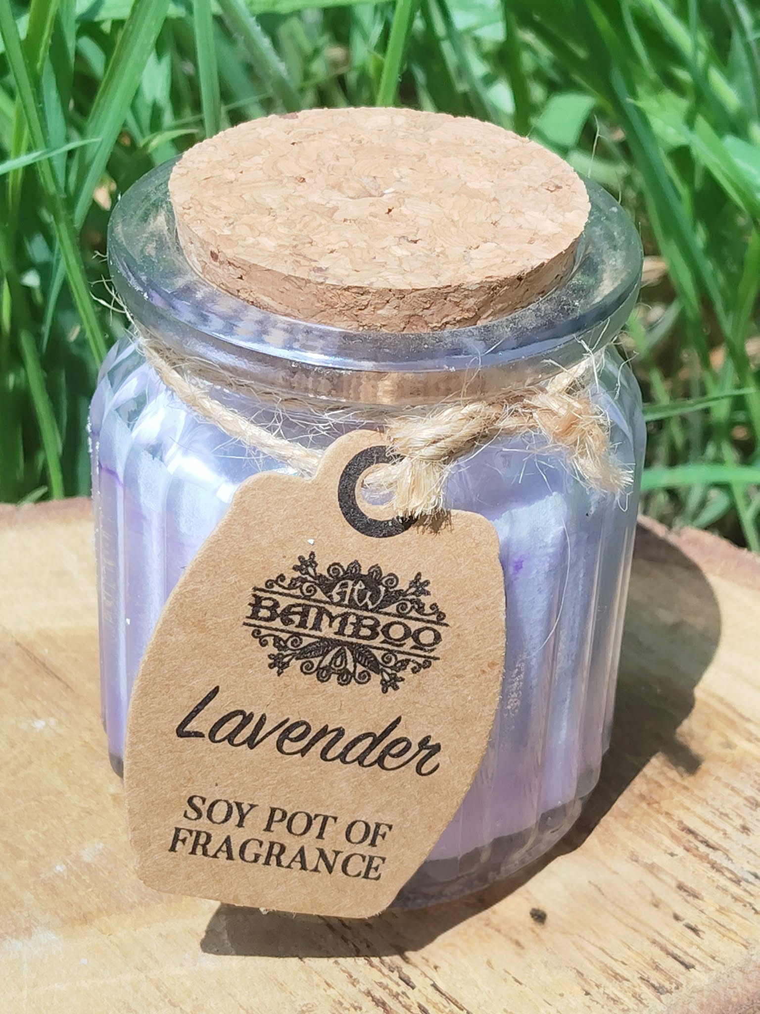 Bougie lavande en pot de verre violet strié avec couvercle en liège et étiquette kraft, photo en extérieur sur support bois, herbe en arrière-plan, ambiance calme et apaisante.