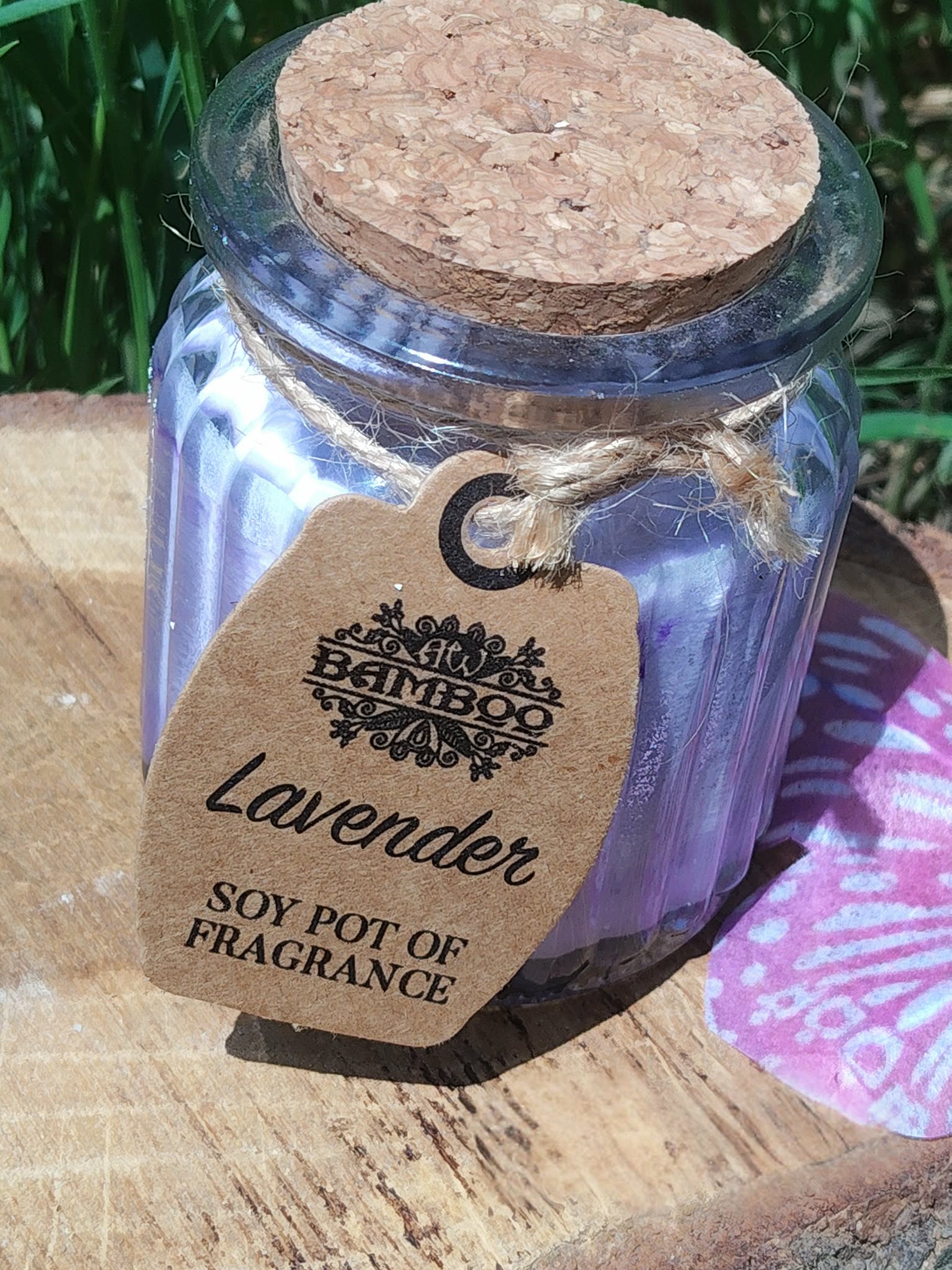Bougie parfumée lavande en pot de verre strié violet avec couvercle en liège, ficelle et étiquette kraft, posée sur planche en bois avec herbe en arrière-plan et papillon violet à côté, ambiance douce, apaisante, détente, sérénité.