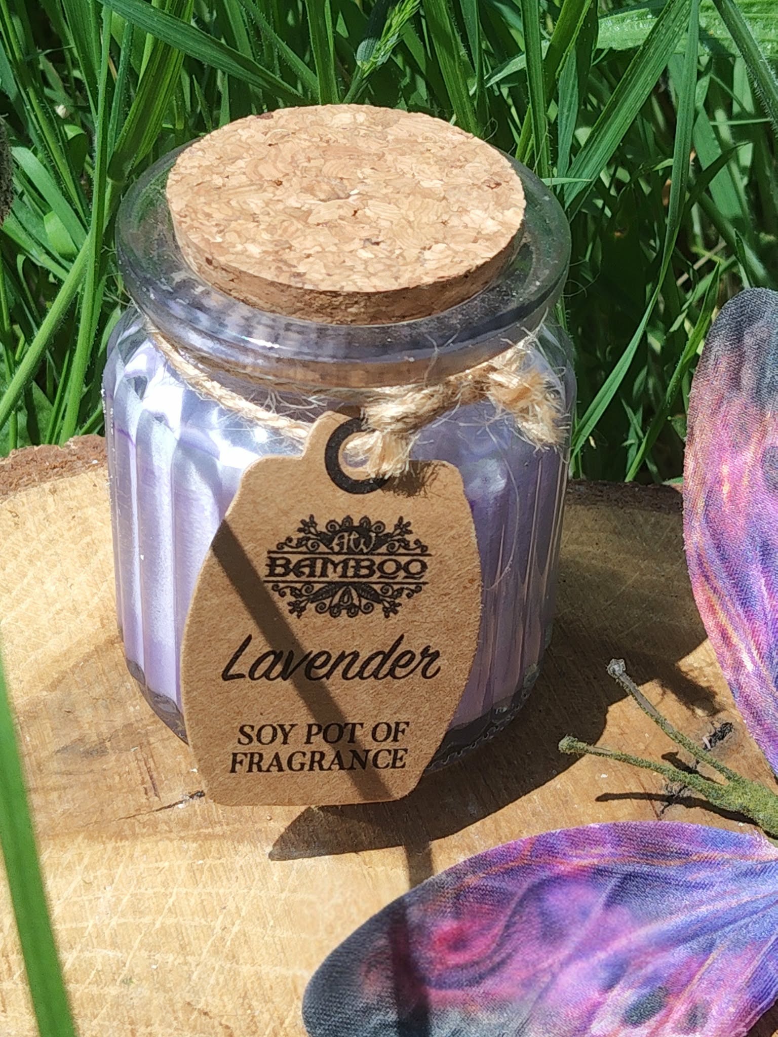 Bougie lavande en pot de verre violet strié avec couvercle en liège et étiquette kraft, posée sur planche en bois en extérieur, herbes en fond, ambiance douce et relaxante.