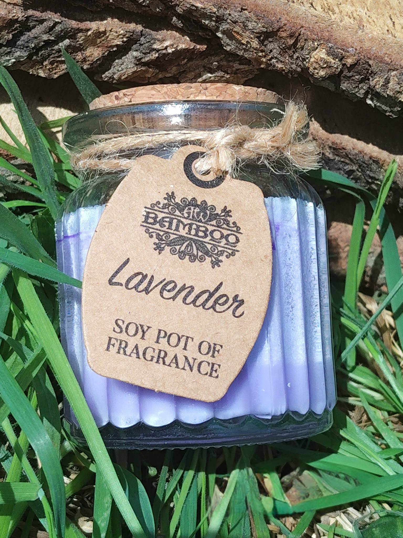 Bougie parfumée lavande en pot de verre strié violet avec couvercle en liège, ficelle et étiquette kraft, posée dans l’herbe en extérieur, lumière naturelle, ambiance fraîche, apaisante, détente, sérénité.