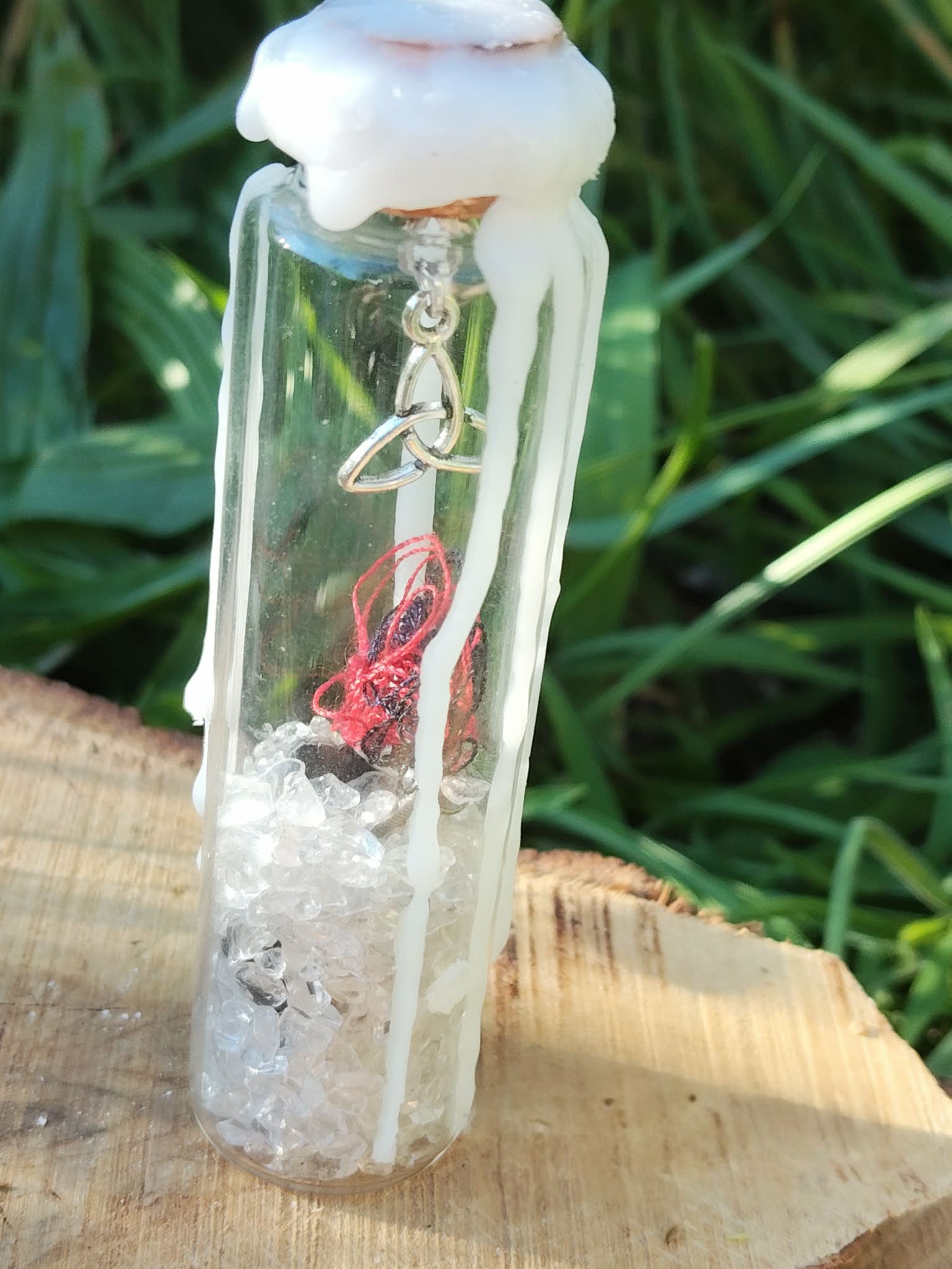 Spell jar en fiole de verre avec cristal de roche, fil rouge, cire blanche et breloque triquetra, posé sur bois en extérieur, ambiance protection et clarté.