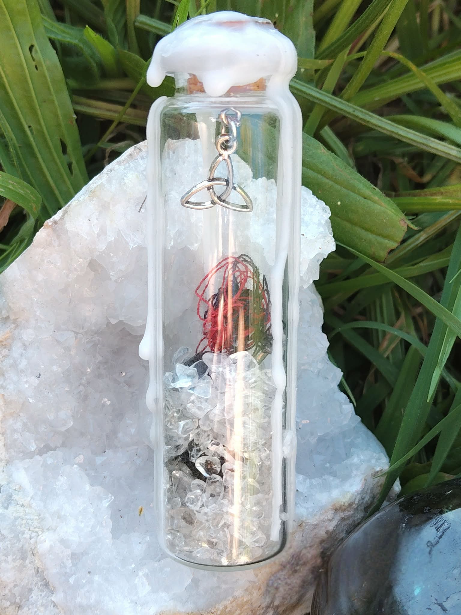 Spell jar en fiole de verre avec cristal de roche, fil rouge et cire blanche, posé sur bois avec coquillage et herbe en fond, ambiance protection et purification.