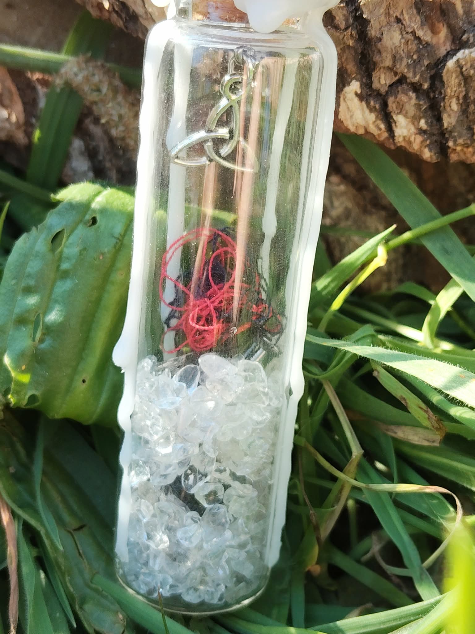 Spell jar en fiole de verre avec cristal de roche, fil rouge, cire blanche et breloque triquetra, photo en extérieur, énergie de protection et clarté.