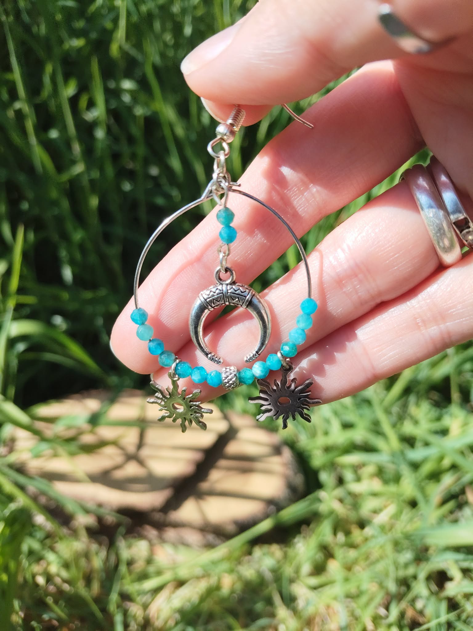 Boucle d’oreille Atlante en apatite bleue création Magic Gaïa tenue entre les doigts, anneau argenté et charms soleil, photo en extérieur sur fond d’herbe et bois, communication, clarté mentale, apaisement, confiance en soi