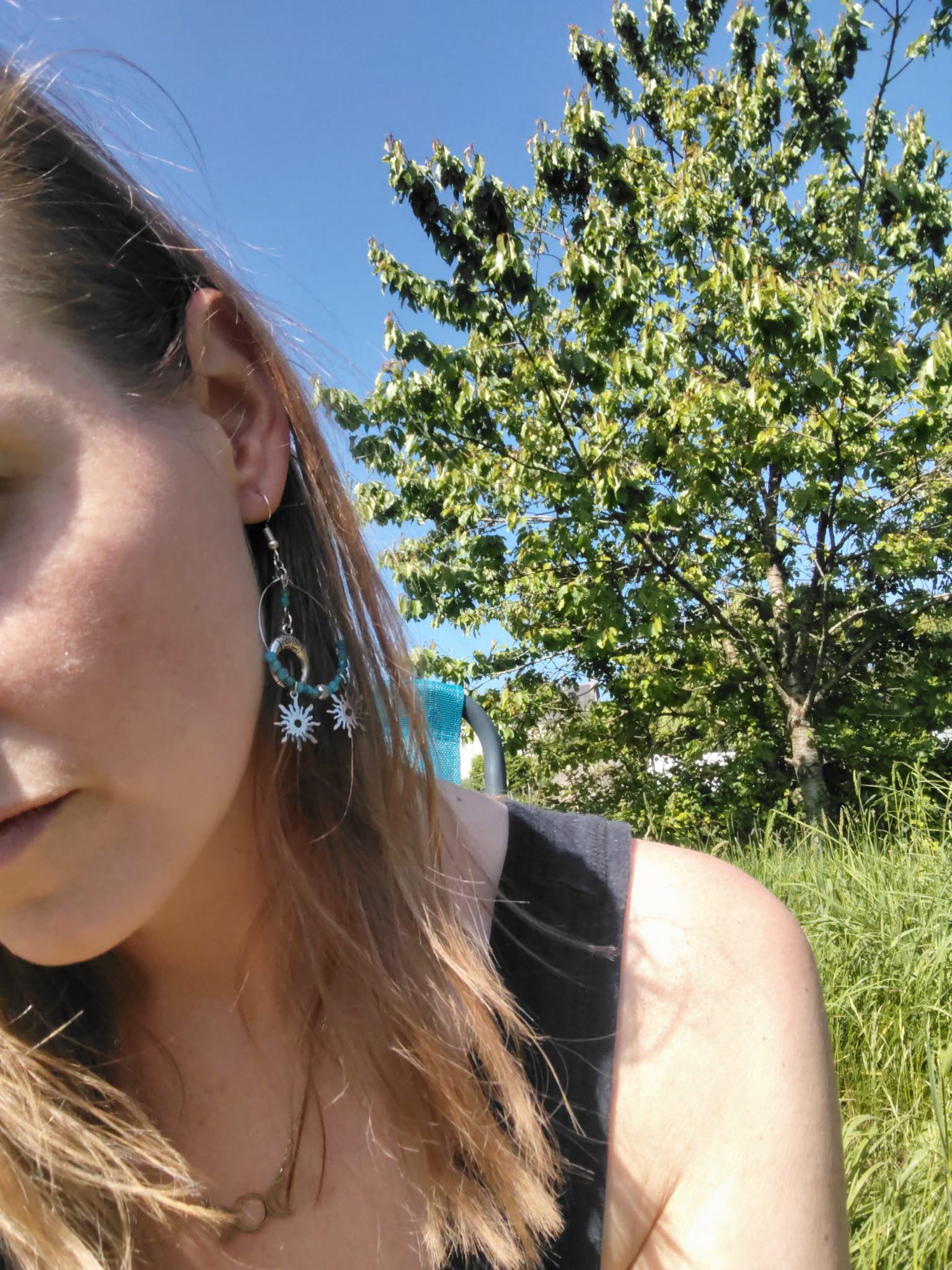 Portrait en extérieur portant une boucle d’oreille Atlante en apatite bleue, création Magic Gaïa, anneau argenté avec charms lune et soleils, fond ciel bleu et arbre. Apaisement, communication, clarté intérieure, confiance douce