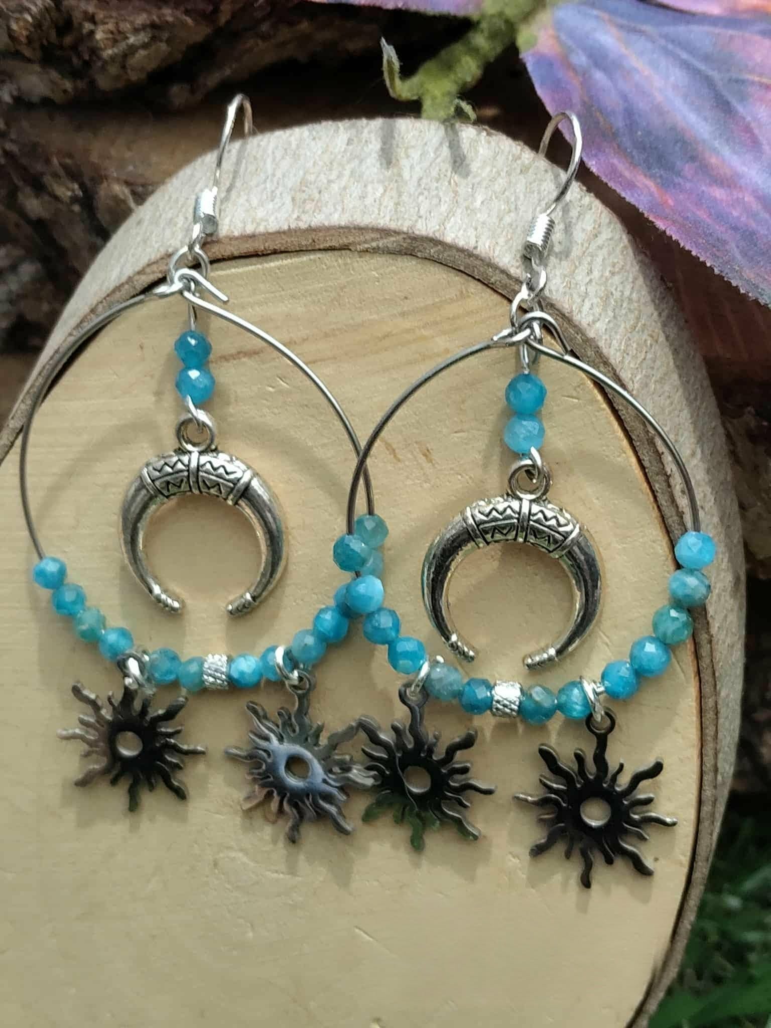Boucles d’oreilles Atlante en apatite bleue, création Magic Gaïa, anneaux argentés avec symbole lune et charms soleil, présentées sur support en bois clair en intérieur. Apaisement, communication, clarté intérieure, confiance douce