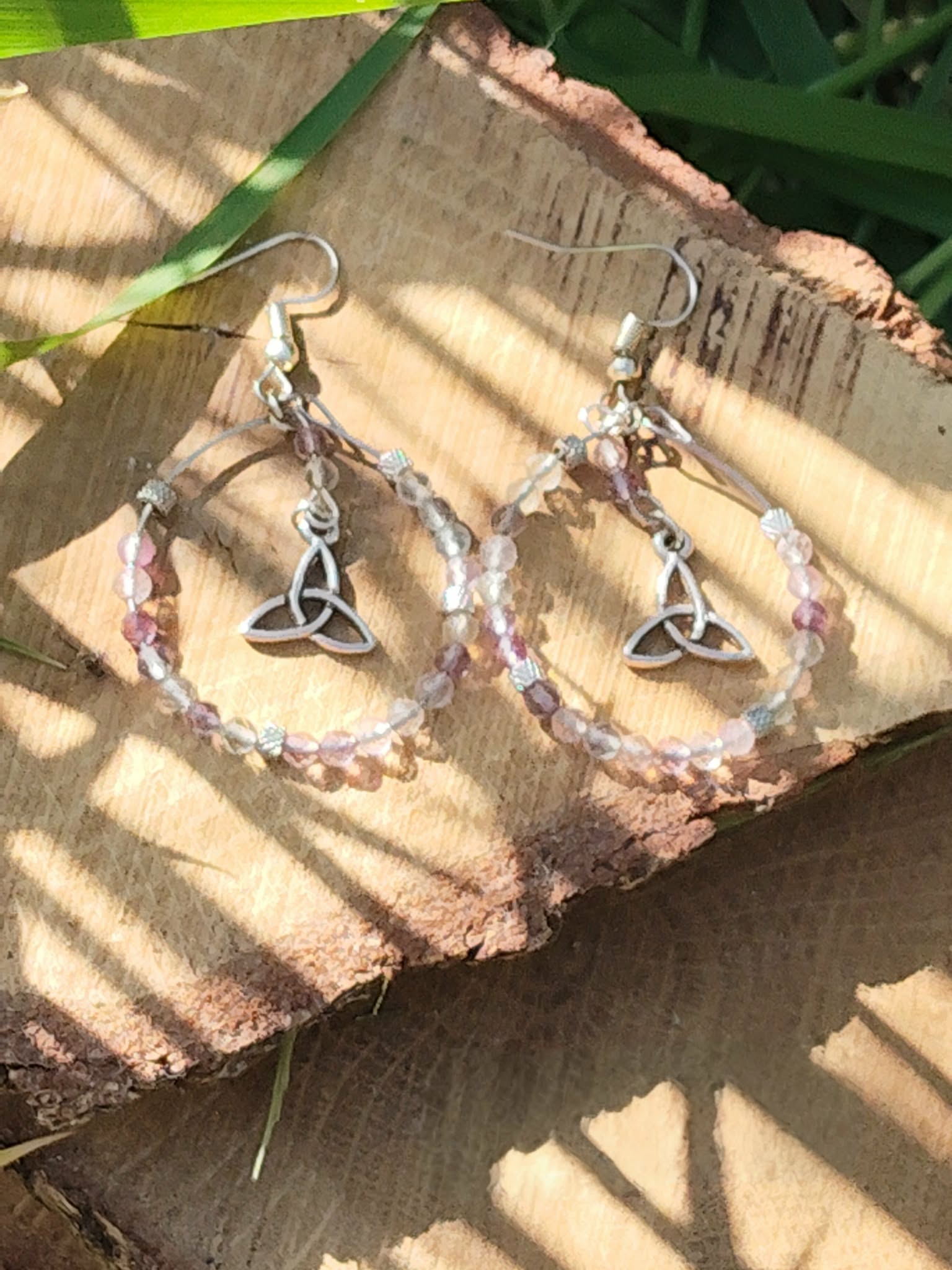 Boucles d’oreilles Hécate en fluorite facettée, création Magic Gaïa, anneaux perlés dégradés et breloques triquetra argentées, posées sur rondin de bois au soleil avec ombres rayées. Clarté, apaisement, intuition