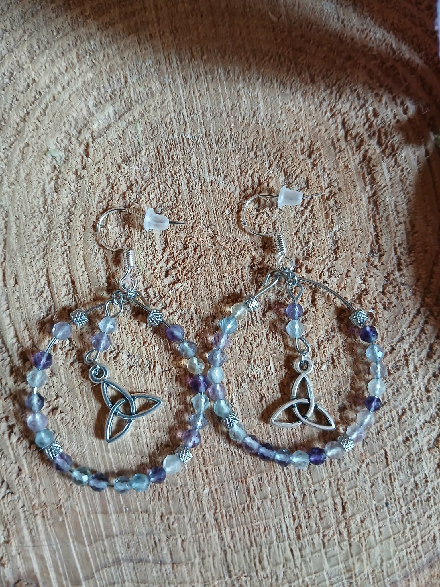 Boucles d’oreilles Hécate en fluorite facettée, création Magic Gaïa, anneaux perlés dégradés et breloques triquetra argentées, posées sur rondin de bois au soleil avec ombres rayées et verdure. Clarté, apaisement, concentration