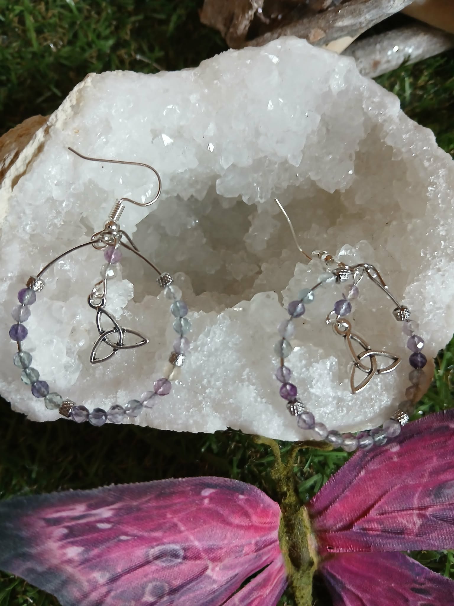 Boucles d’oreilles Hécate en fluorite facettée, création Magic Gaïa, anneaux perlés dégradés et triquetra argentée, posées sur quartz blanc avec ailes de papillon roses en décor. Clarté, apaisement, intuition