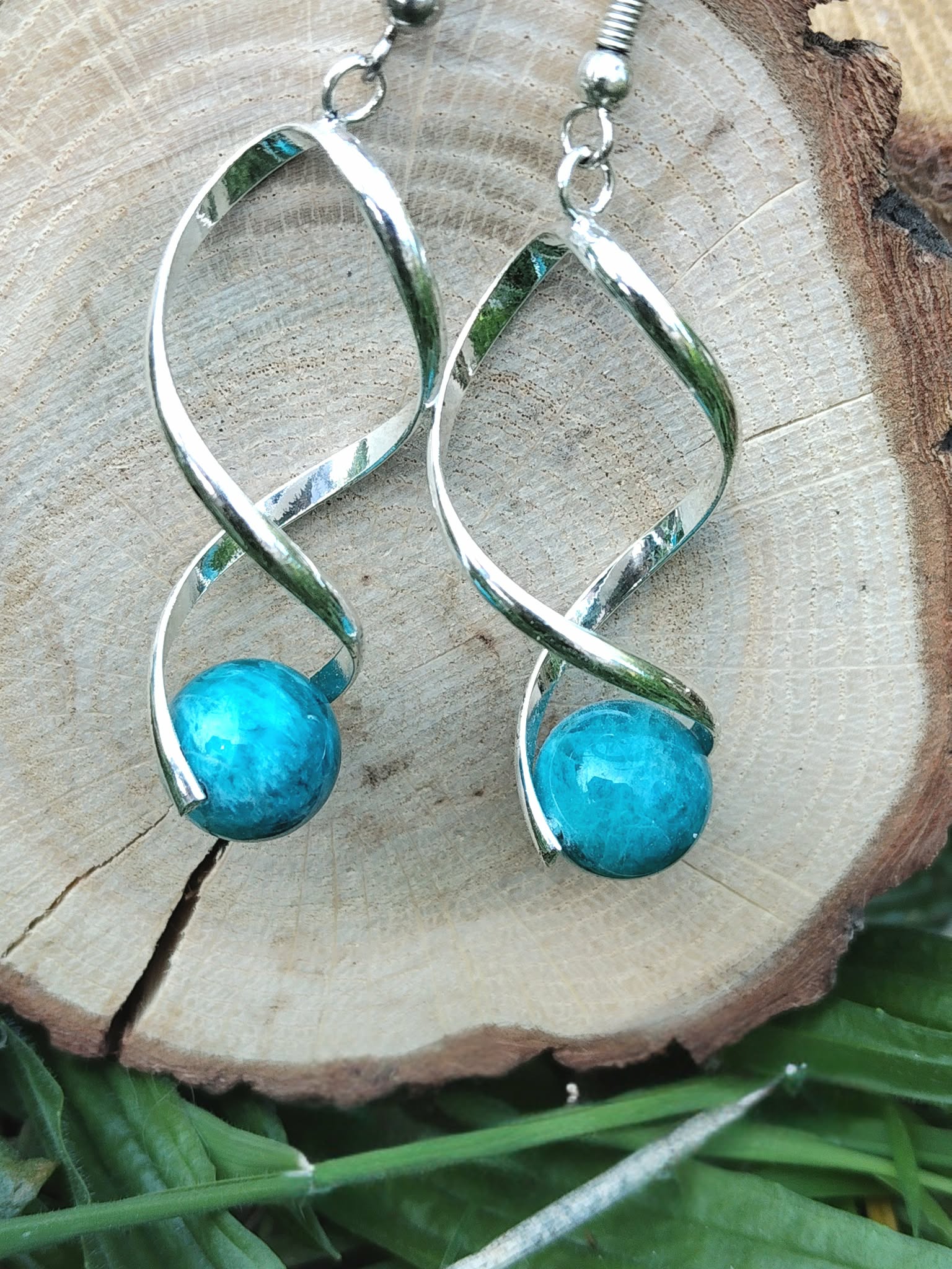 Boucles d’oreilles spirales argentées avec perles d’apatite bleue posées sur rondin de bois et décor végétal, clarté, motivation, confiance.
