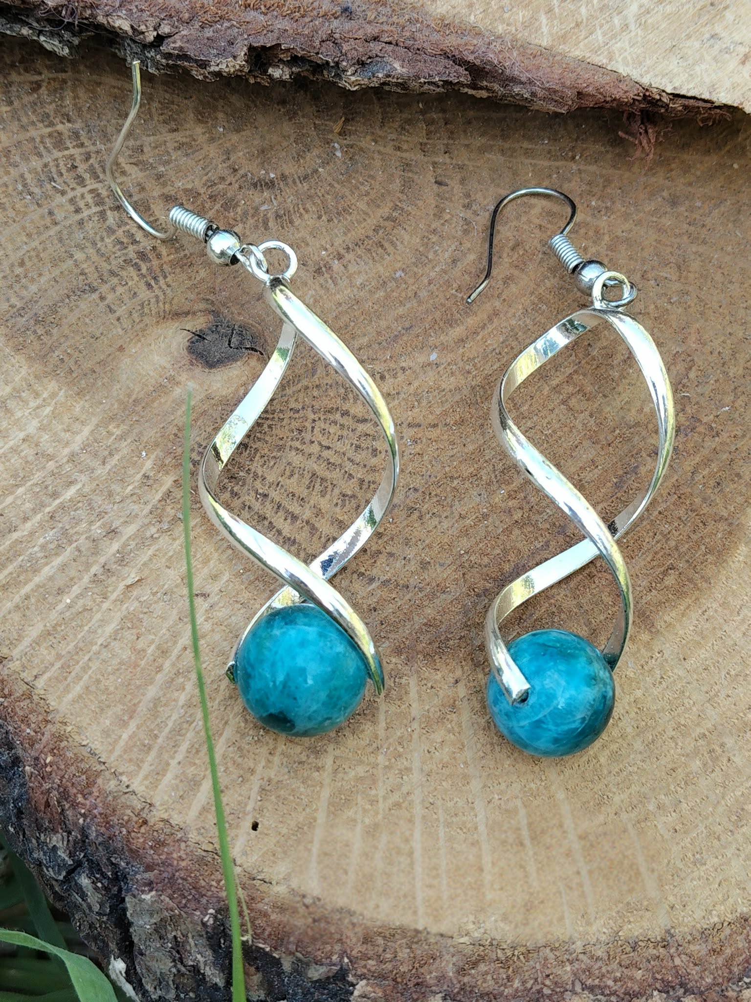 Boucles d’oreilles spirales argentées avec perles d’apatite bleue posées sur rondin de bois et décor végétal, clarté, motivation, confiance.