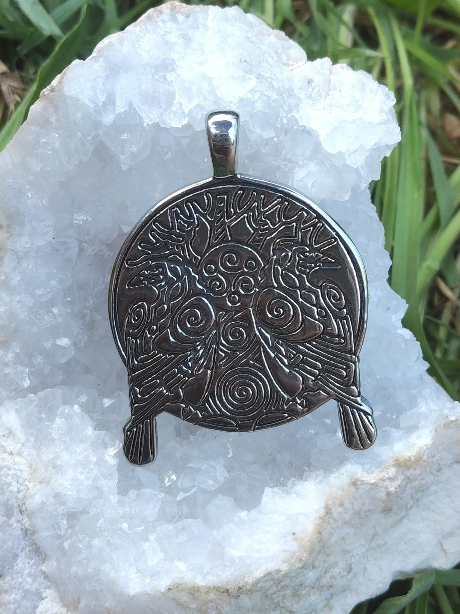 Pendentif viking en métal noirci avec arbre sacré et corbeaux en relief, posé sur géode blanche scintillante et feuillage vert