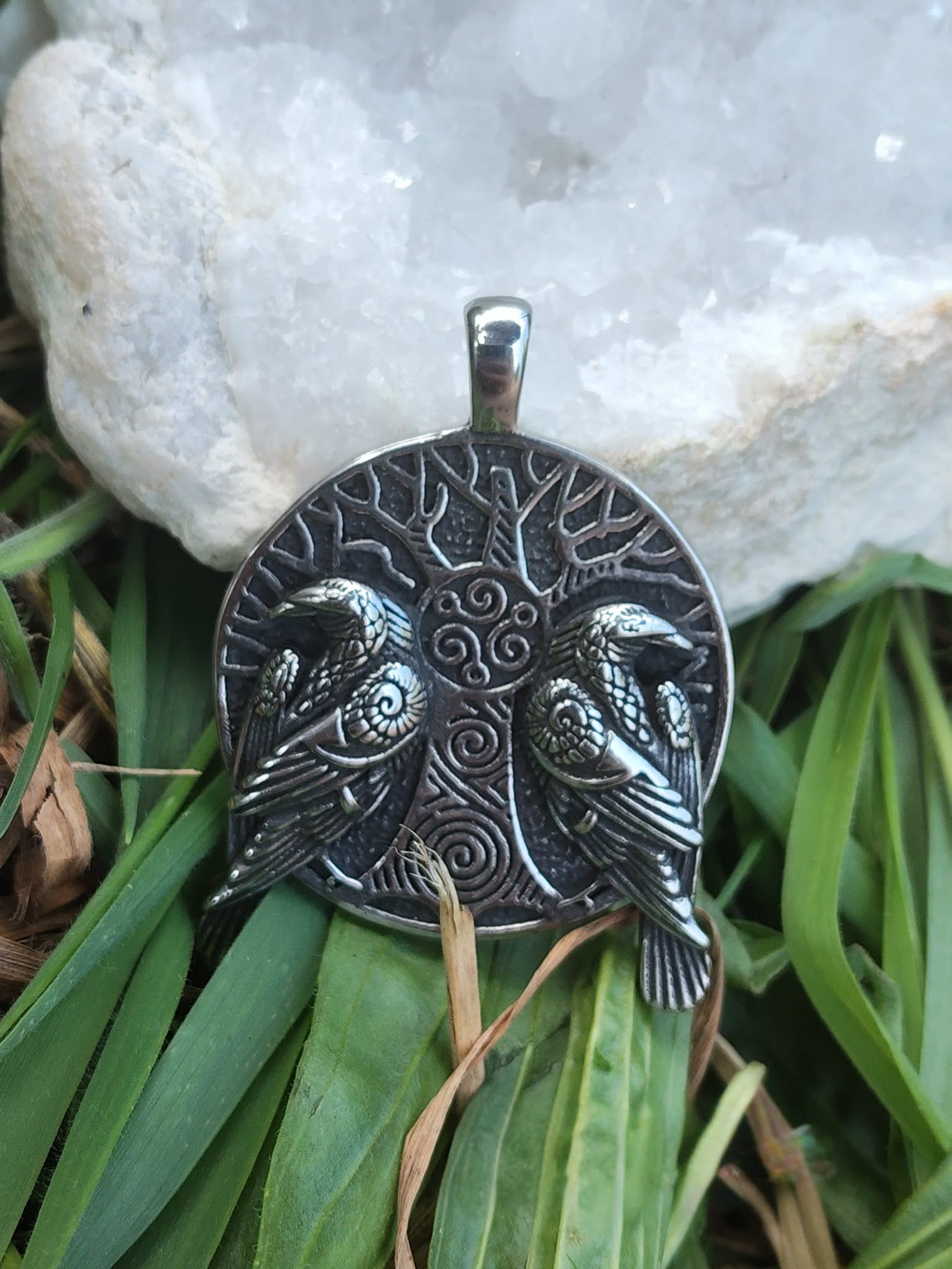 Pendentif viking verso argenté noirci avec gravure arbre sacré et spirales, posé devant mousse, feuillage vert et amas de quartz