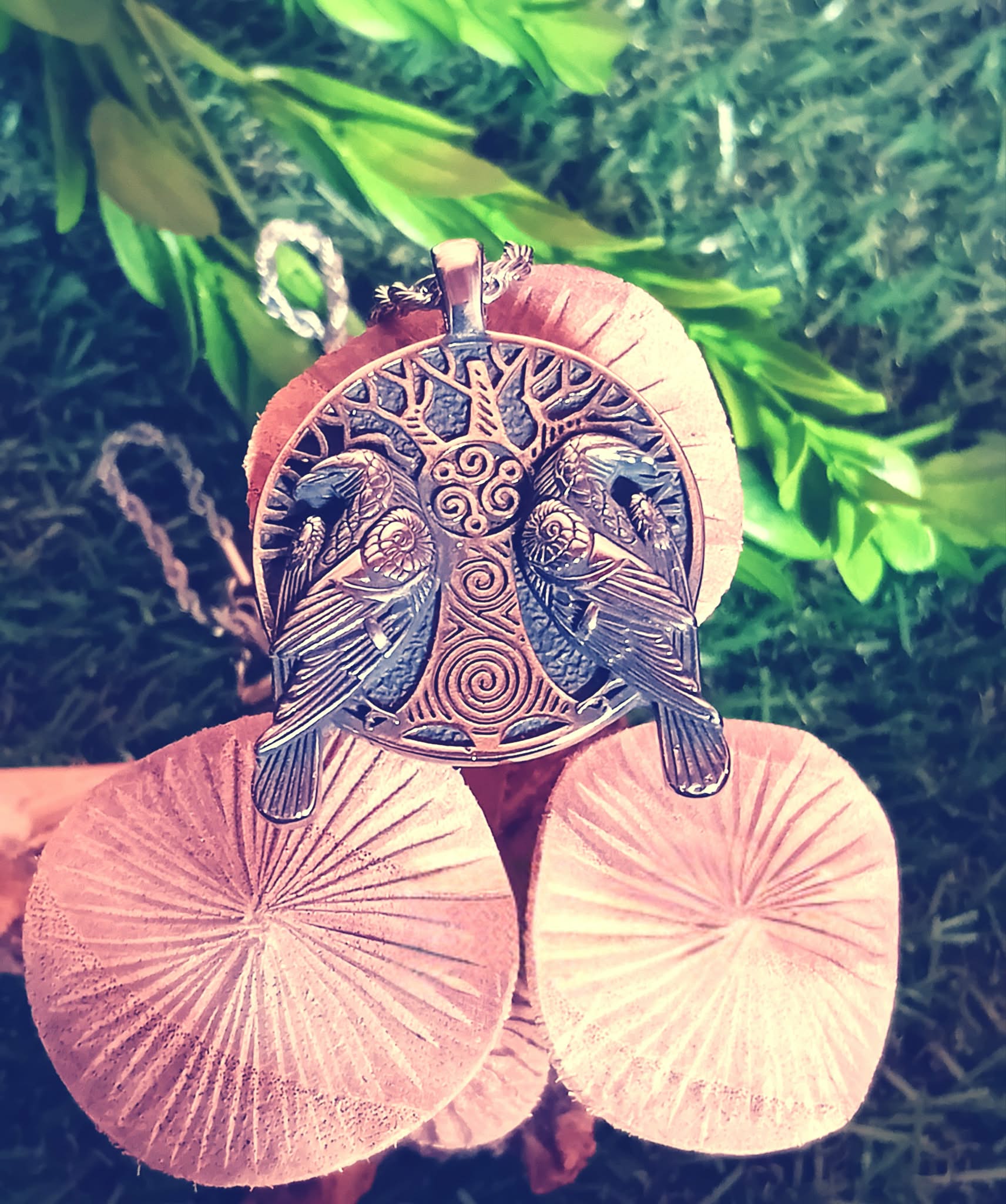 Pendentif viking en métal argenté vieilli avec arbre sacré et deux corbeaux, posé sur champignon en bois miel avec herbes vertes floues