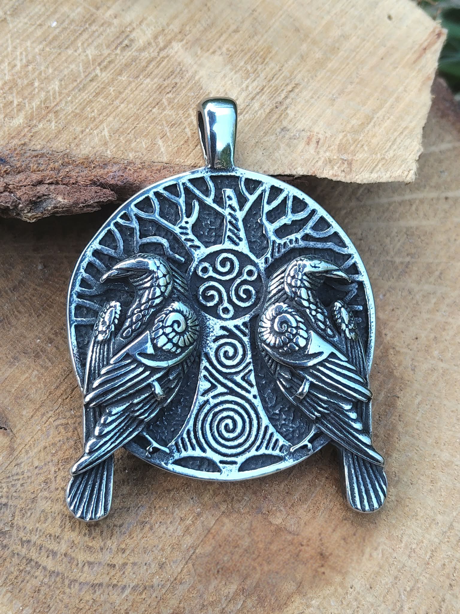 Gros plan sur pendentif viking argenté noirci avec arbre sacré, doubles corbeaux et spirales, posé sur  bois ton miel