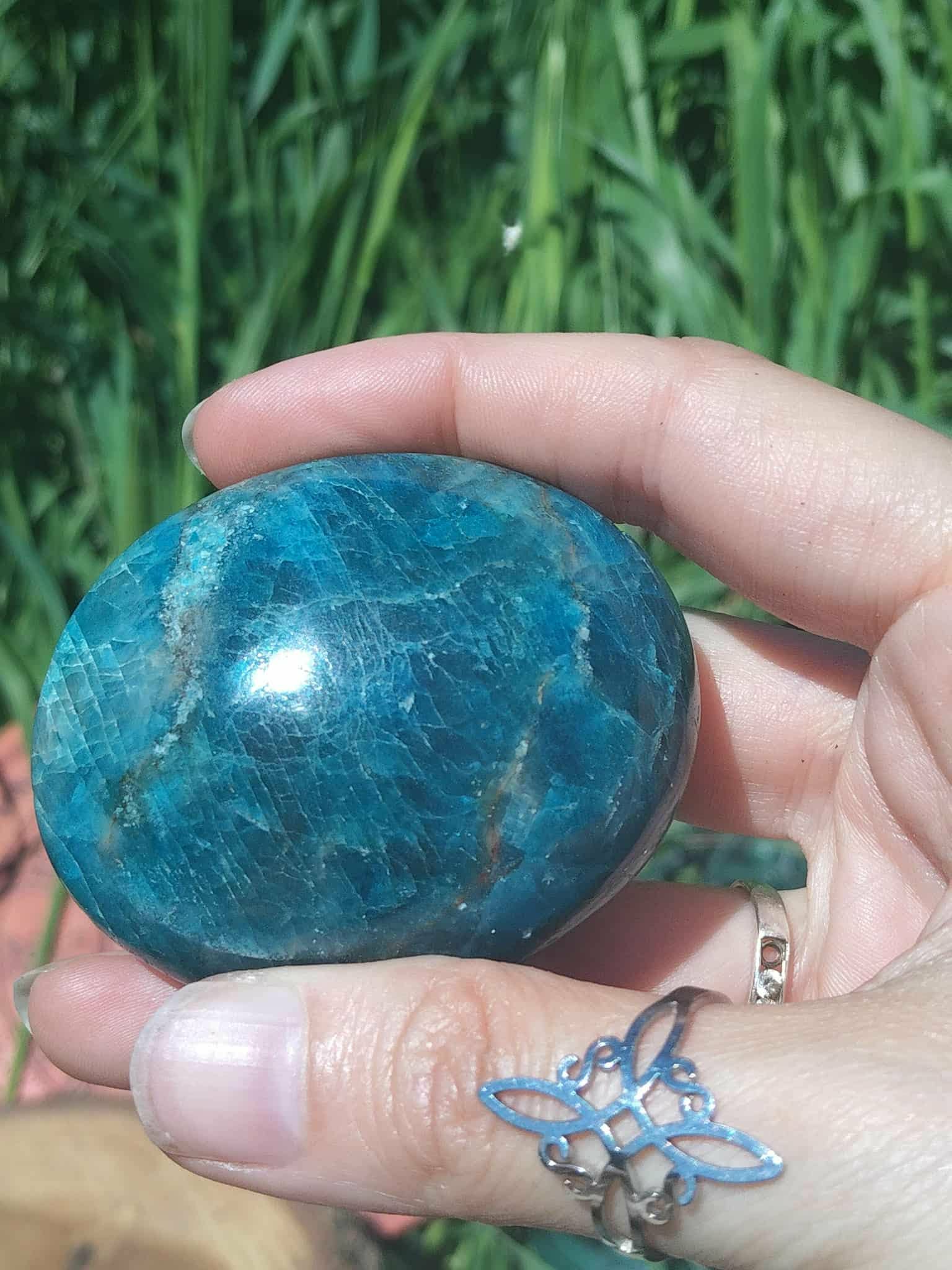 Pierre roulée d’apatite bleue poli bleu lagon marbré, tenue entre les doigts devant des herbes vertes, pierre naturelle liée à l’authenticité, au recentrage et à la fluidité d’expression.