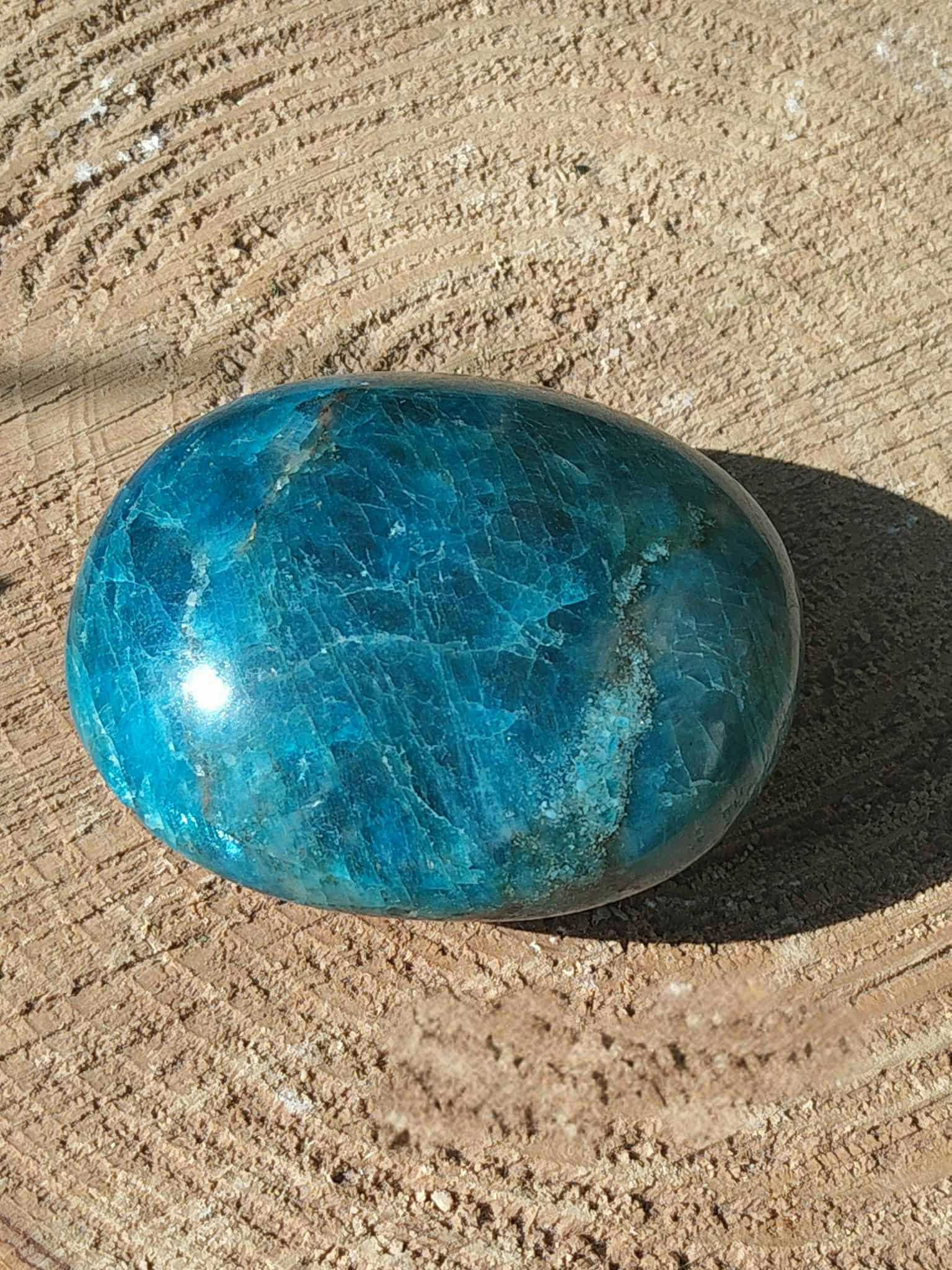 Pierre roulée d’apatite bleue aux nuances bleu lagon marbrées, posée sur support beige texturé avec ombre portée, pierre naturelle associée au discernement, au calme du mental et au dynamisme doux.